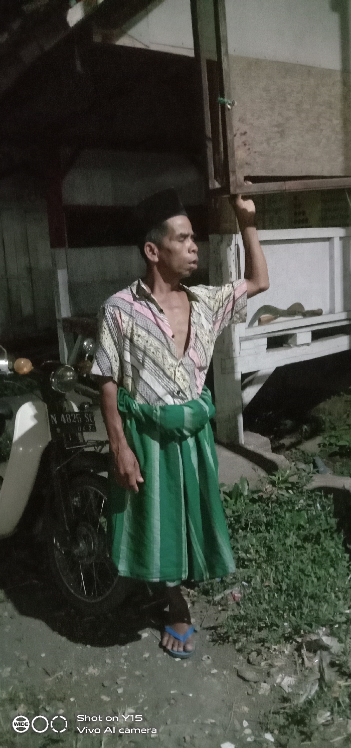 An adult male stands outdoors at night in Ranugedang, Indonesia. He is wearing a black songkok, an unbuttoned patterned short-sleeved shirt featuring diagonal stripes of pink, white, and beige, a green and lighter green striped sarong, and blue flip-flops. His right arm is raised, hand gripping an overhead dark wooden beam. His left arm hangs by his side. He gazes to the right, with his mouth slightly ajar. A light-colored motorcycle with license plate 'N 4825 SG' is partially visible to the left. In the background, positioned to the right of the man, a curved blade tool with a light-colored handle rests on a white wooden structure. The ground consists of dirt with patches of green vegetation. The scene is illuminated by an artificial light source, creating shadows.