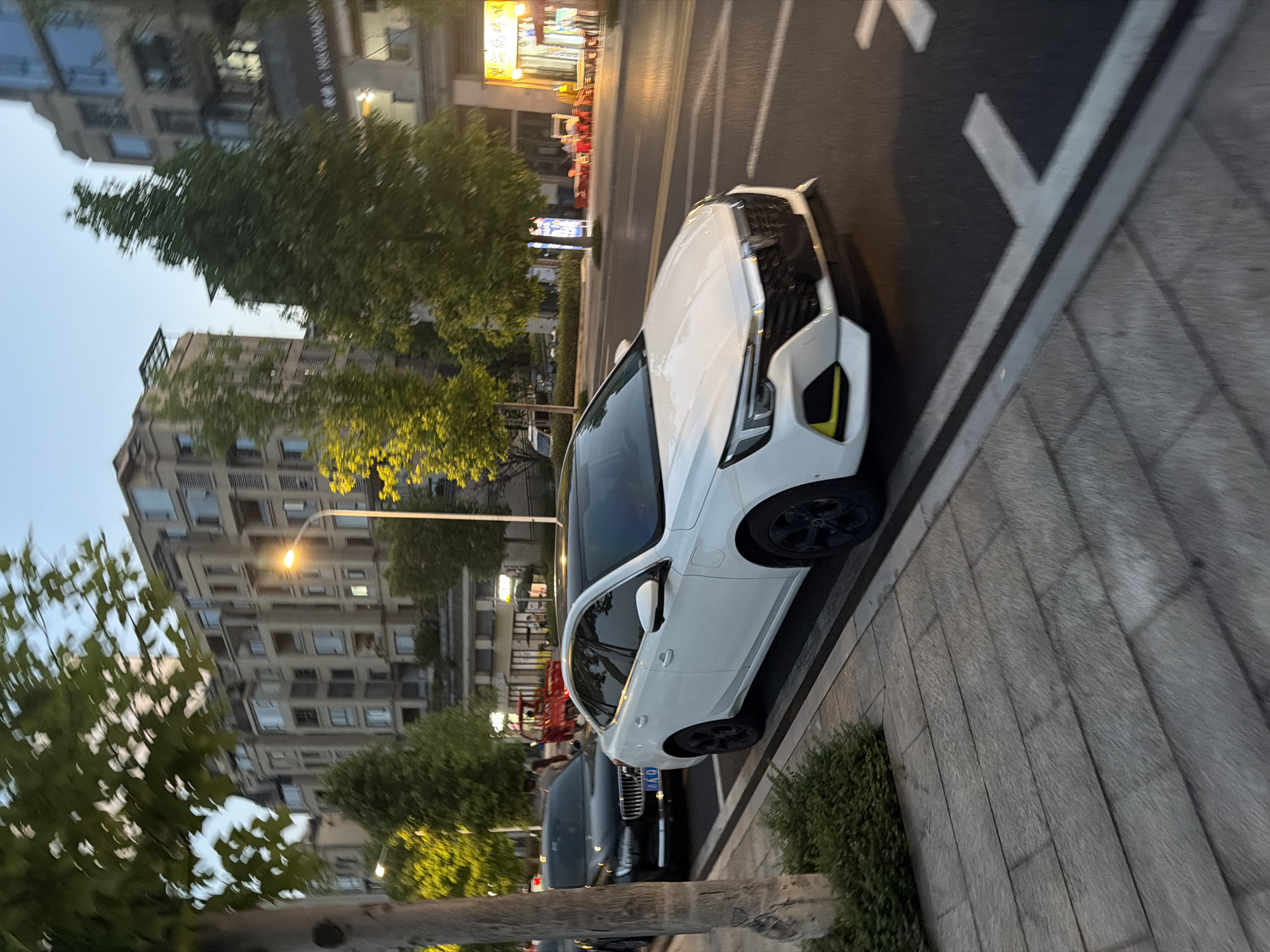 The image shows a street scene in Jiangjin, China. In the foreground, a white sedan is parked in a designated parking spot on the right side of the road. The car has a black roof and black wheels with yellow accents on the front bumper. To the left of the white sedan, a black sedan is also parked. Behind the cars, a sidewalk made of rectangular stone pavers is visible, with a small hedge in front of it.
The street itself is paved with asphalt and marked with white lines. In the background, there are multi-story buildings with balconies and windows. Trees with green foliage line the street, and a streetlamp is illuminated. Further in the background, there are glimpses of shops with yellow awnings and red outdoor seating. A red three-wheeled vehicle is also visible in the distance. The sky is a soft blue, suggesting it is either early morning or late evening.