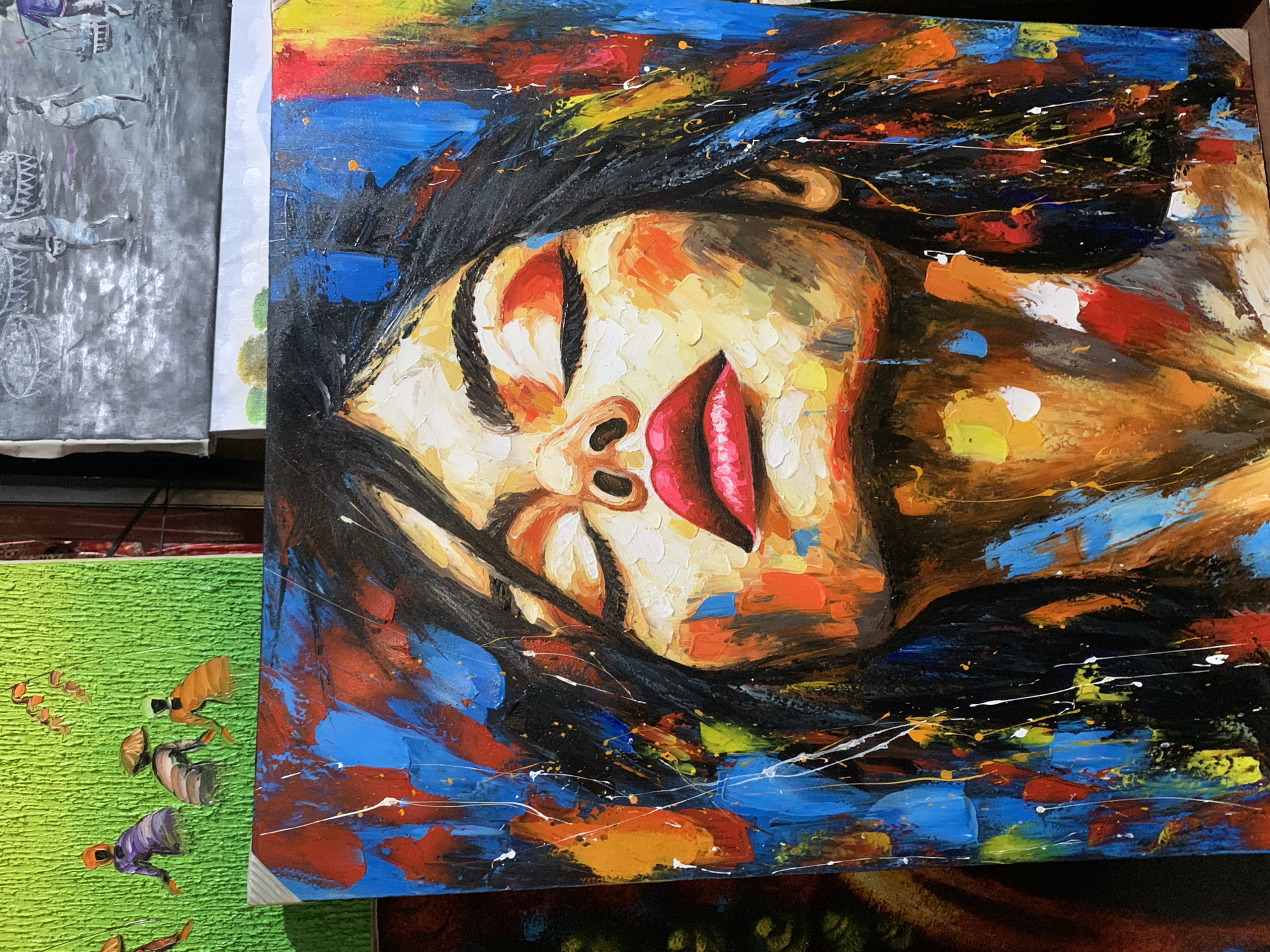 The image shows a vibrant, abstract painting of a woman with her eyes closed, seemingly asleep or in repose. The artwork is characterized by thick impasto paint application and a vivid color palette including blues, reds, yellows, oranges, blacks, and whites. The woman's face is rendered in warm tones, while her hair and the background feature bold strokes of contrasting colors. The painting style is expressive and energetic, with splatters and visible brushstrokes adding to the dynamic feel of the piece.
The painting appears to be part of a display or for sale in a location in the city of Ubud District, Indonesia, near other paintings, including a gray painting of horses and carts. Additionally, a green textured painting is seen with small figures of people with hats.