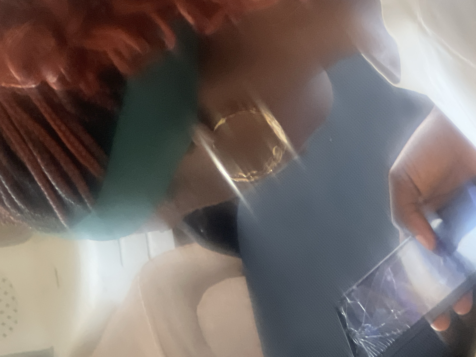 The image shows a close-up of a person's profile, focusing on their head and hands. The person has dark skin and braided hair, some of which is red. They are wearing a teal headband and large gold hoop earrings. Their right hand is holding a smartphone with a cracked screen, and they appear to be looking at it. The background is blurred but suggests an indoor setting with soft lighting and a lighter-colored surface, possibly a sofa or chair. The overall scene is casual and personal, with no discernible elements that would definitively place it in Jos, Nigeria.