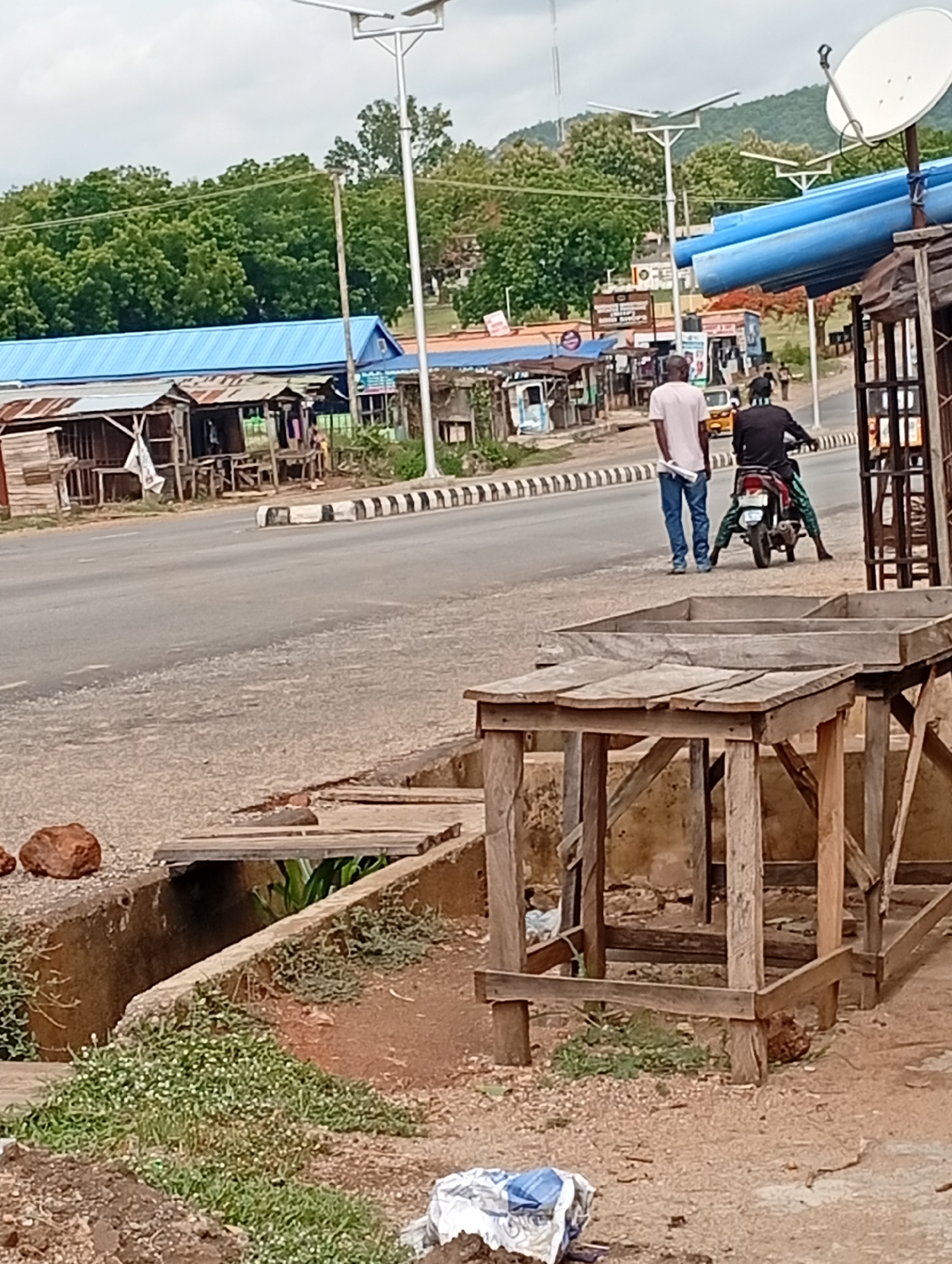 The image shows a street scene in Adankolo, Nigeria. A paved road runs horizontally through the middle of the frame, with a black and white striped barrier on one side. Several small, weathered shops or stalls line the far side of the road, some with blue roofs. Two men are on the road, one standing in a white shirt and jeans, and the other sitting on a green and silver motorcycle. In the foreground, there are wooden structures, possibly tables or stalls, and a ditch with a wooden plank across it. The ground is dirt, with sparse vegetation and some trash. Streetlights are visible along the road. In the background, there are green trees and a hill. The sky is overcast.