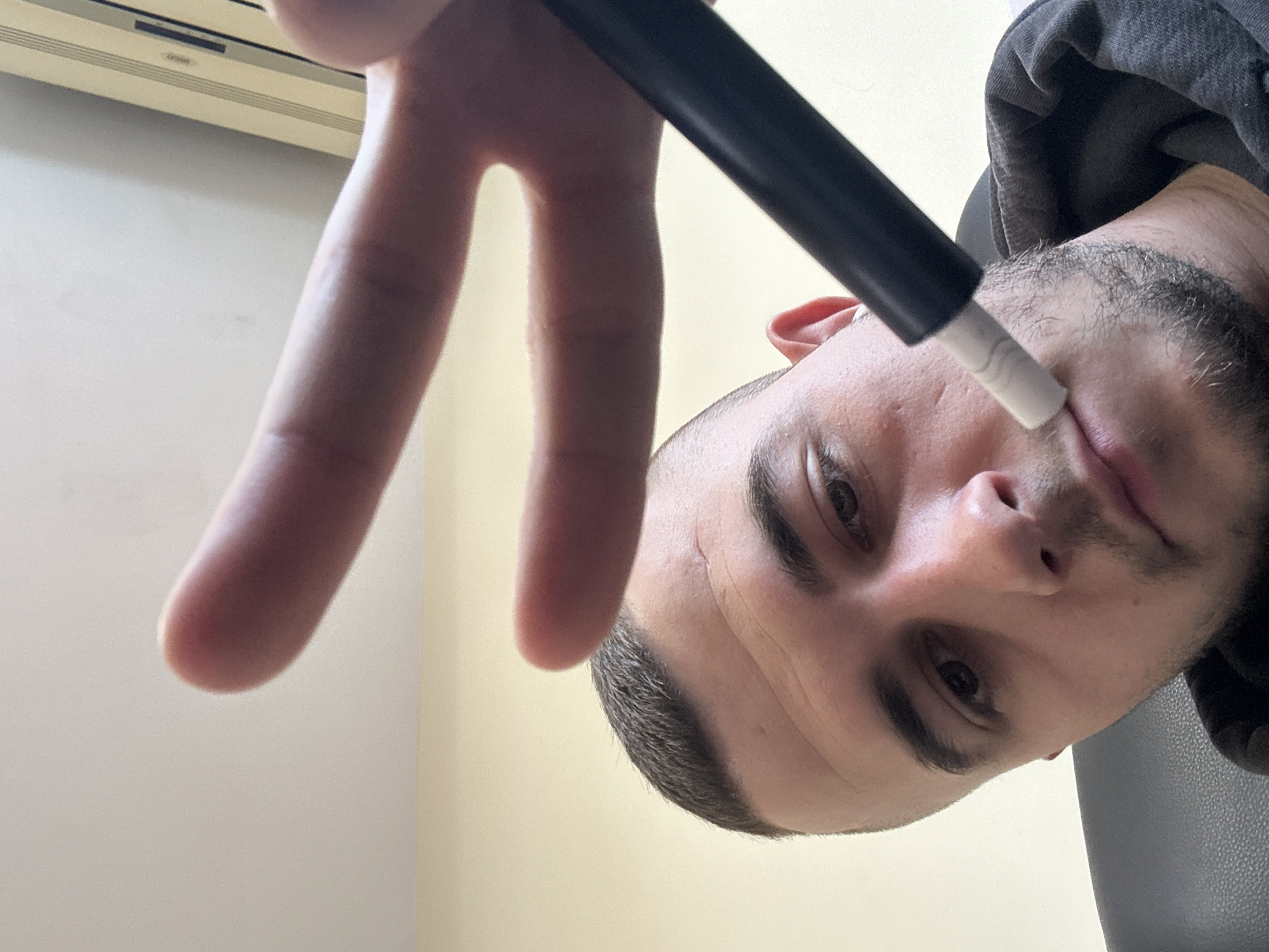 A close-up, angled shot features a man's face and hand. His left hand is held up, fingers spread in a "peace" sign. In his right hand, he holds a black wand with a white tip near his nose. He is wearing a dark hooded sweatshirt. The background shows a cream-colored wall and a white air conditioning unit. There are no indications or hints in the image to confirm a specific location in Plovdiv, Bulgaria.