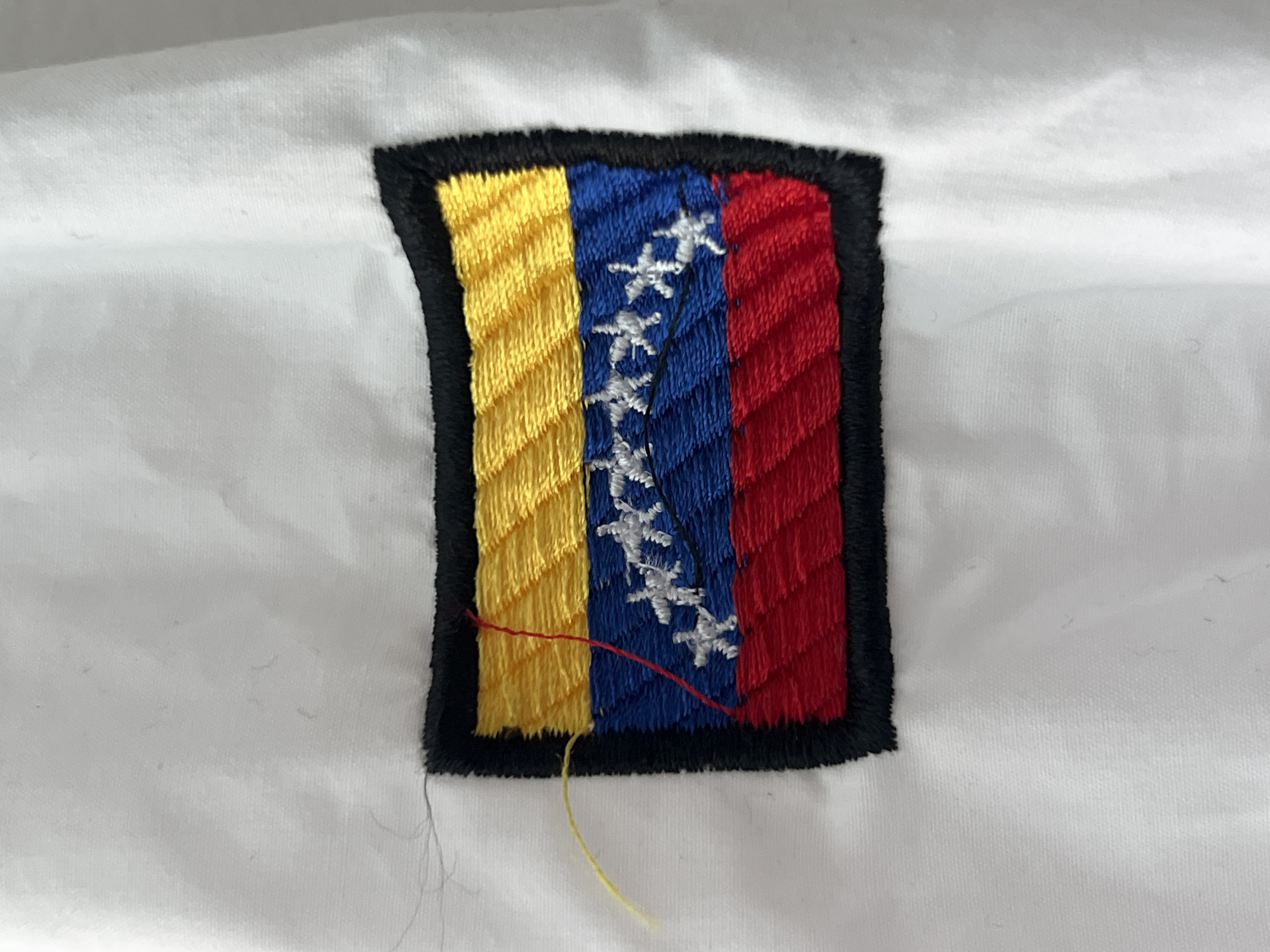 The image presents a close-up view of an embroidered patch affixed to a white fabric background. The patch is rectangular with slightly rounded corners, outlined by a thick, solid black embroidered border.
The central design of the patch resembles a flag, composed of three vertical stripes: yellow on the left, blue in the middle, and red on the right. The yellow and red stripes feature diagonal embroidery stitches, while the blue stripe uses vertical stitches. Positioned vertically along the blue stripe are eight white, five-pointed stars, also embroidered. These stars have a slightly irregular, handcrafted appearance.
From the bottom edge of the patch, two loose threads are visible: one red thread extends from the lower part of the blue stripe, and one yellow thread hangs from the bottom-left corner of the black border. This detail might indicate an unfinished work or loose stitching.
The white background material appears to be a piece of fabric, possibly a garment, exhibiting some subtle creases and a soft texture. The lighting is bright and even, highlighting the colors and the textured nature of the embroidery. There are no people, discernible activities, or visible text in the image. The flag design strongly resembles that of Venezuela.