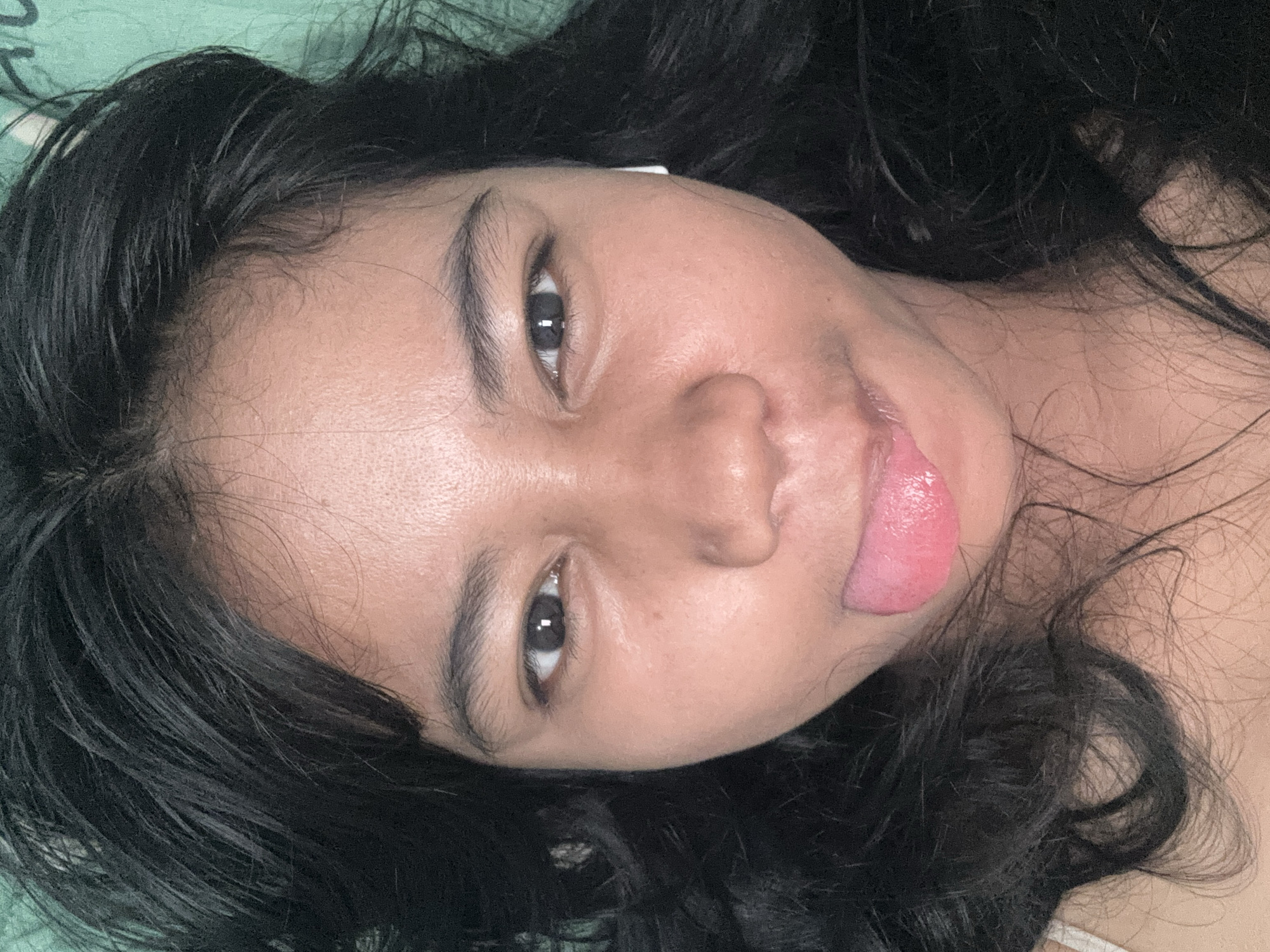 A close-up, over-the-shoulder, selfie-style photograph shows a young Indonesian woman with long, dark hair lying down with her tongue sticking out. She is wearing a white tank top and an earbud in her right ear. The background is a green textured surface. The image was taken indoors in North Jakarta, Indonesia.