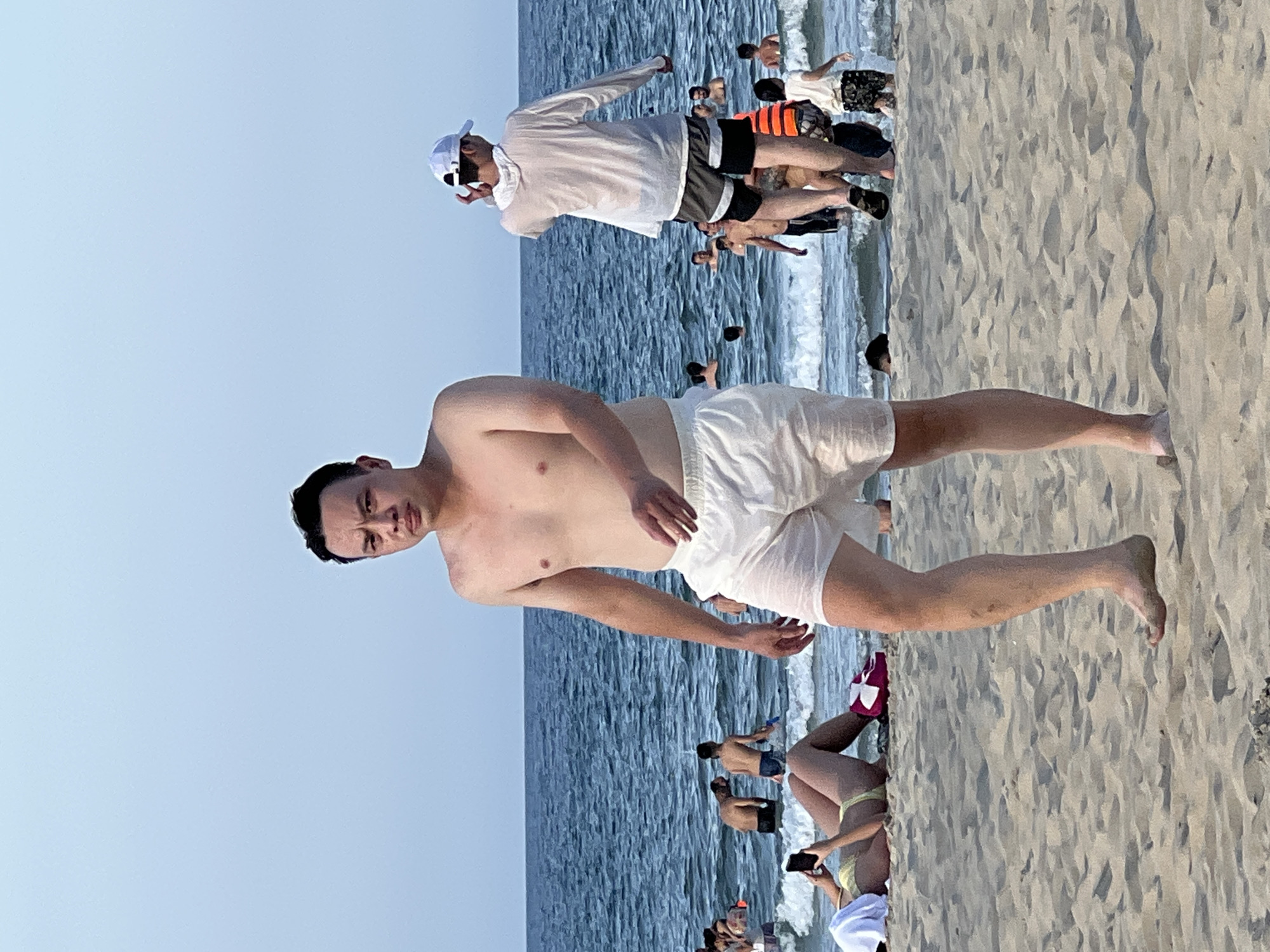 A group of people are at a beach in Đà Nẵng, Vietnam. The main subject is a man walking towards the camera in white shorts. In the background, other people are in the ocean, some swimming and some standing in the shallow water. One person is lying on the sand looking at their phone. The sky is clear and blue, and the ocean is a deep blue. The sand is light brown. The image is taken during the day.