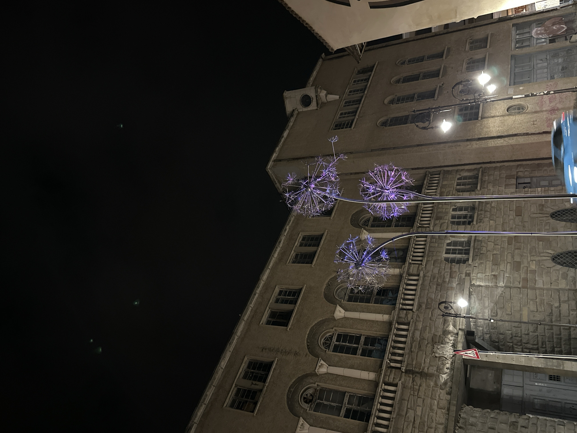 The image shows a low-angle nighttime view of a tall building with multiple floors. The building has a mix of rectangular and arched windows, and its facade appears to be made of stone or concrete. Three illuminated spherical decorative elements resembling dandelion seed heads are suspended in front of the building. A street lamp with a light on is also visible, along with a traffic sign. The sky is dark and there are faint light spots in the dark sky, possibly from city lights. The building's architecture and decorations suggest it is located in a European city. It is NOT located in the city of Veliko Tarnovo, Bulgaria.