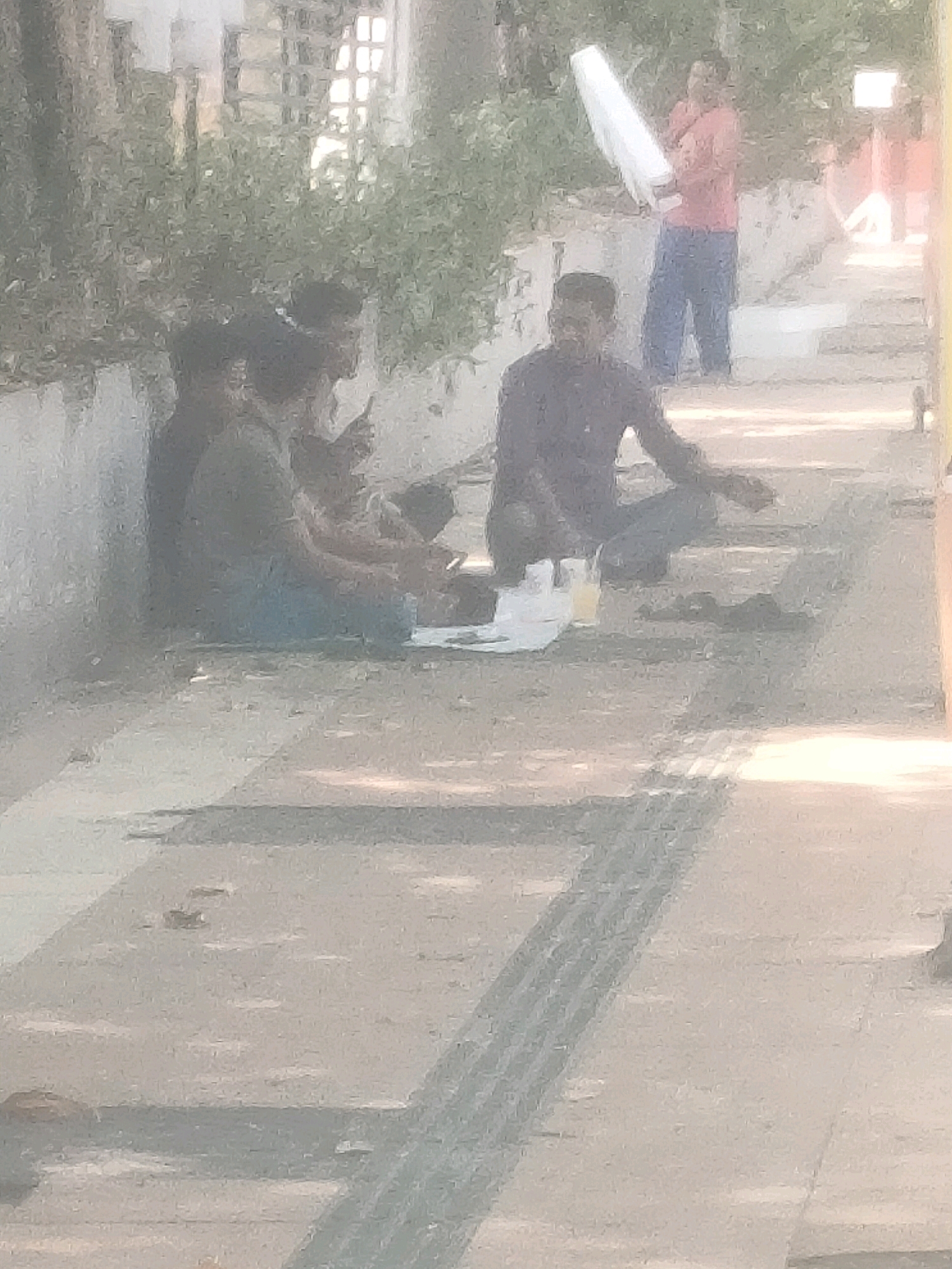 In Surabaya, Indonesia, a group of men are gathered on a sidewalk next to a street. The foreground shows a paved surface with textured lines, indicating a pedestrian pathway. In the mid-ground, several individuals are seated on the ground, some appearing to be eating from containers placed on a white sheet. One man, wearing a dark shirt, is actively engaged with the food. To the right, another man in a dark shirt is also seated. Further in the background, a person in a red shirt holds a long, white object, possibly a rolled-up poster or a piece of equipment. The environment is outdoors, with foliage visible behind the seated individuals and buildings in the distance, suggesting an urban setting. The lighting indicates it is daytime, with shadows cast on the ground.