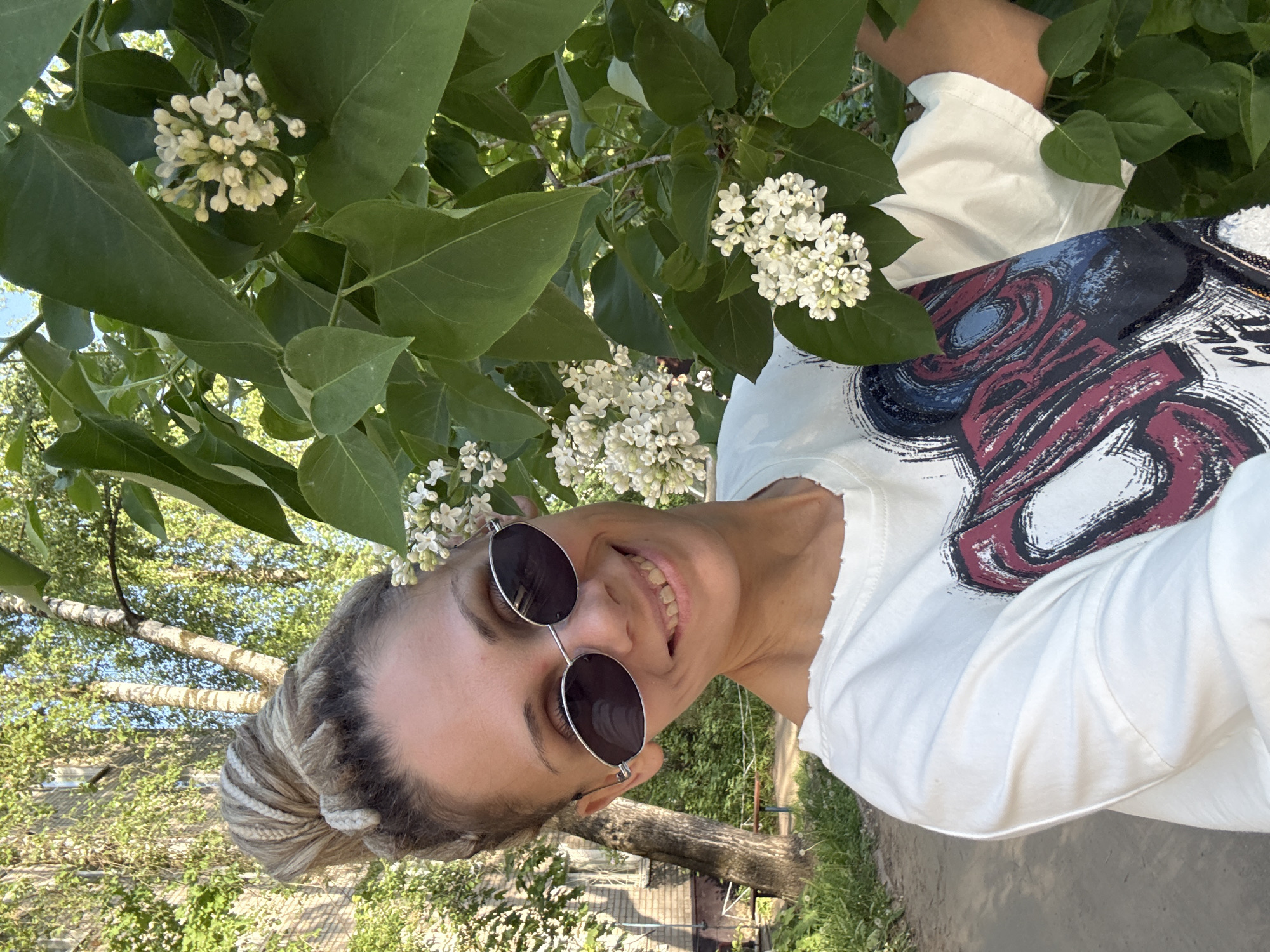 A woman with blonde braids stands near white lilacs in Rusanovo, Russia. She wears round sunglasses and a white patterned t-shirt. The background includes foliage, trees, and a glimpse of buildings.
