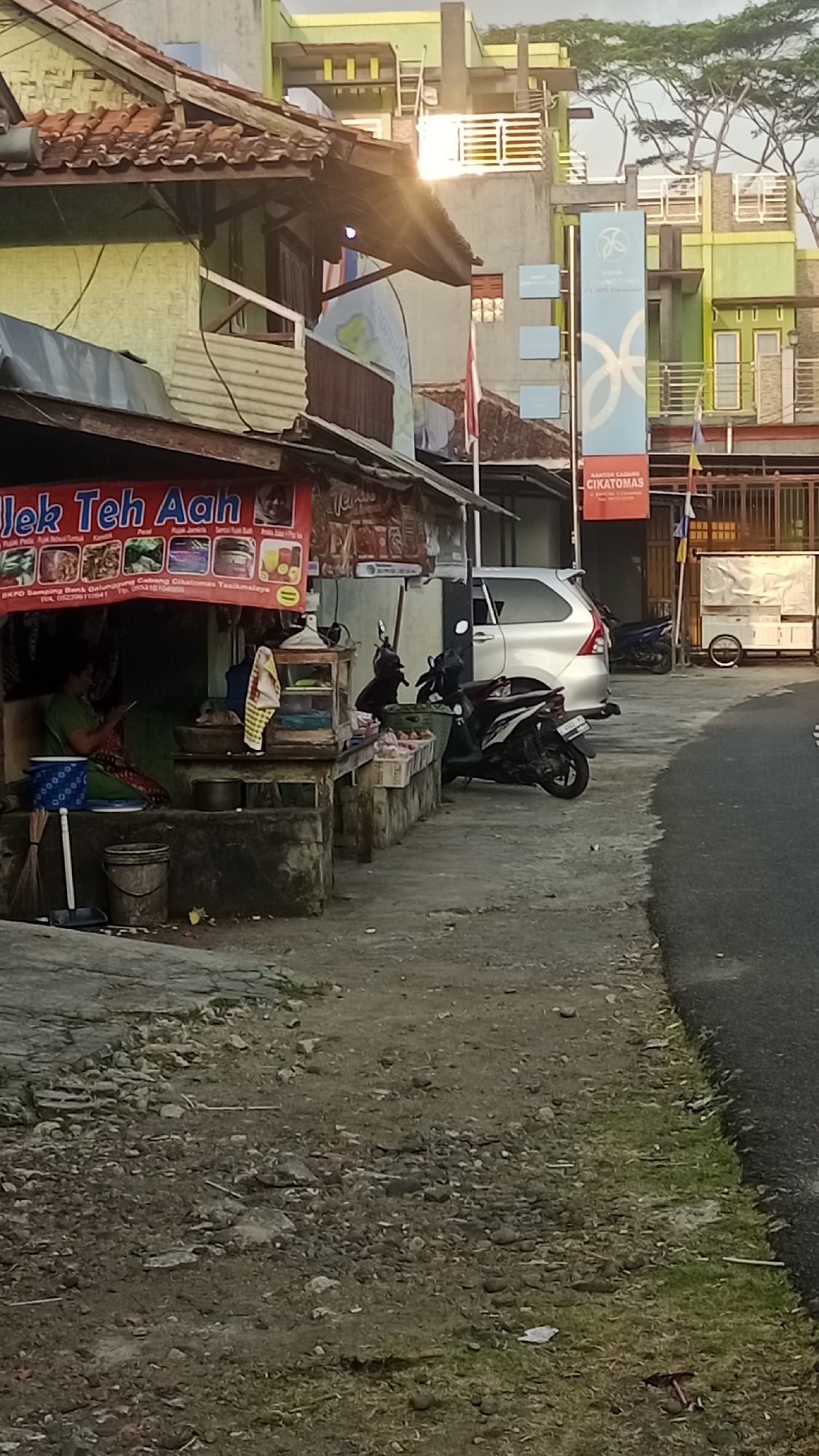 The image depicts a street scene in Pakemitan, Indonesia, likely in the late afternoon or early evening given the lighting. The foreground shows a rough, gravelly ground with scattered stones and some patches of dry grass. A paved pathway or road runs along the right side of the image, with its edge bordered by more grass and debris.
To the left, a small roadside stall is visible. A woman, wearing a green shirt and a patterned skirt, is seated behind the stall, appearing to use a mobile phone. She is positioned next to a stack of what look like woven baskets or containers. In front of her, there are various items on display, including a display case with shelves and items on top, possibly food or drinks. A broom and a bucket are also present near her.
Behind the stall, a red banner with Indonesian text, "JLEK TEH AAH," is prominently displayed, suggesting it is a food or beverage vendor. Further along the stall, there are more banners with text and images. A motorcycle is parked directly in front of the stall.
Further back in the scene, a silver car is parked on the side of the road. Adjacent to the car, another motorcycle is parked. A white food cart with a canopy is also visible further down the street.
The background features several buildings with a distinctive architectural style, featuring light green and pale yellow facades, with multiple stories and balconies. One building has a tall, vertical banner with a blue logo and text, including "CIKATOMAS." An Indonesian flag is also visible. Trees with sparse foliage are present on top of some of the buildings. The overall impression is of a bustling, everyday street scene in a residential or commercial area.
