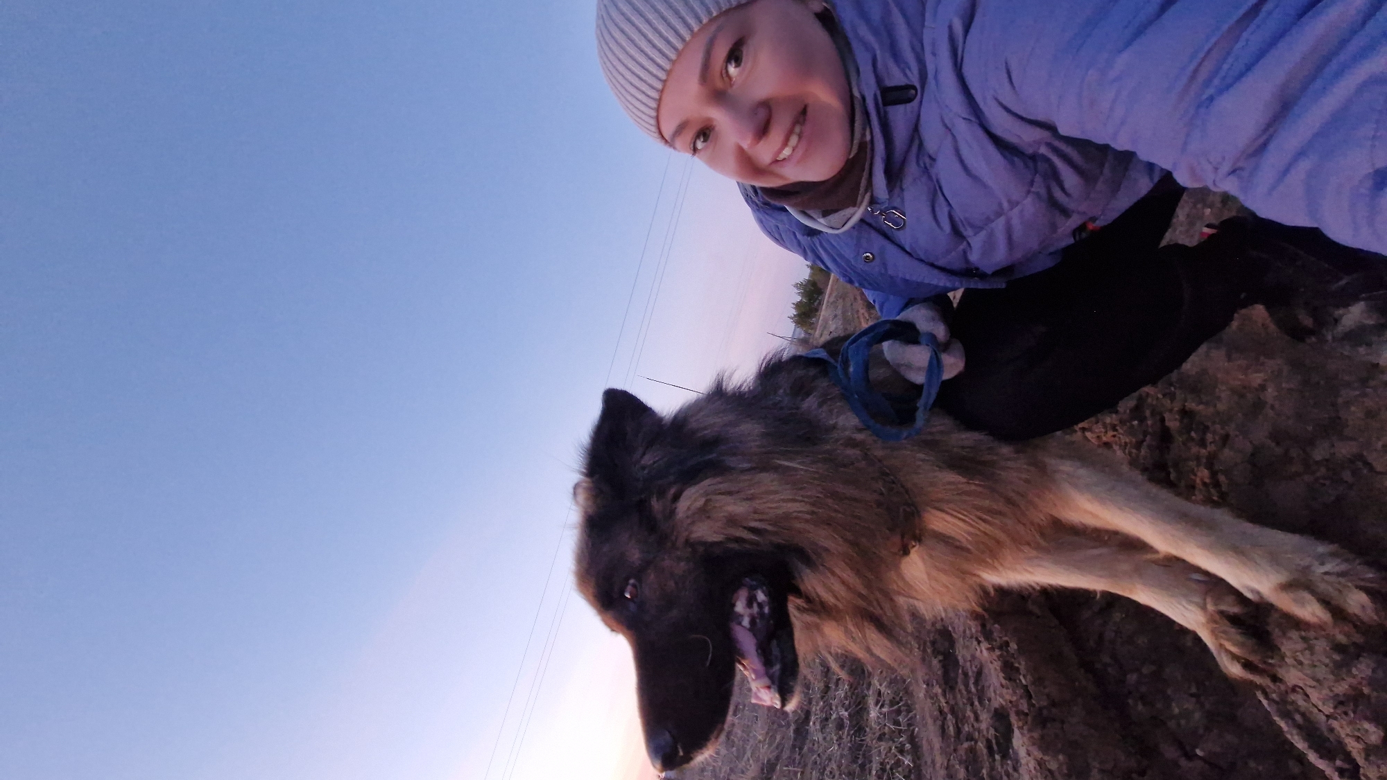 The image captures a close-up selfie of a woman and her large dog outdoors in Otchor, Russia, likely during either sunrise or sunset. The woman is positioned in the upper right, smiling widely towards the camera. She wears a light grey ribbed beanie and a light purple or blue quilted jacket. Her left hand, gloved, holds a blue leash connected to the dog.
The dog, a large, fluffy breed with brown and black fur, occupies the lower and central part of the frame. Its mouth is open, tongue visible, suggesting it might be panting or in an expressive state. It appears to be looking towards the camera alongside its owner.
The background consists primarily of a clear sky, displaying a gradient from a deep blue at the top to a softer, lighter blue with hints of pinkish-purple closer to the horizon. Thin overhead lines are faintly visible in the mid-ground. The lower right portion of the image shows dry, uneven ground, possibly dirt or sparse vegetation. The overall mood conveys a moment of happy companionship between the woman and her pet during an outing in an open, natural environment. No visible text is present.