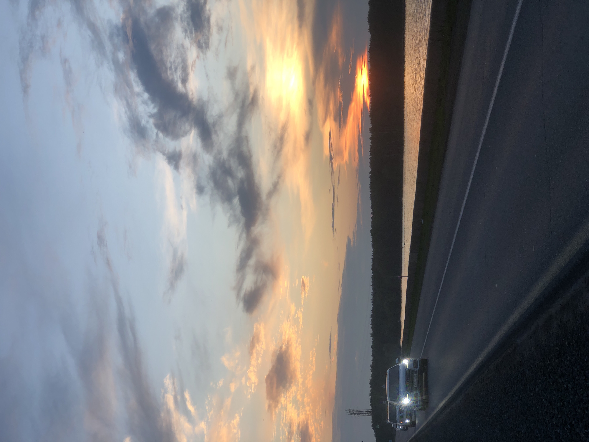 A photograph captures a roadside scene at dusk in Chaikovsky, Russia. The sky is filled with a gradient of colors, transitioning from pale blue at the top to a warm orange and yellow near the horizon, with scattered clouds. A body of water, reflecting the sky's colors, runs parallel to a dark, tree-lined shore. In the foreground, a paved road is visible, with a dark shoulder to the left. Two cars are driving on the road; the one in the foreground is a silver SUV with its headlights on, and another vehicle is behind it. A power line tower can be seen in the distance on the left side of the image.