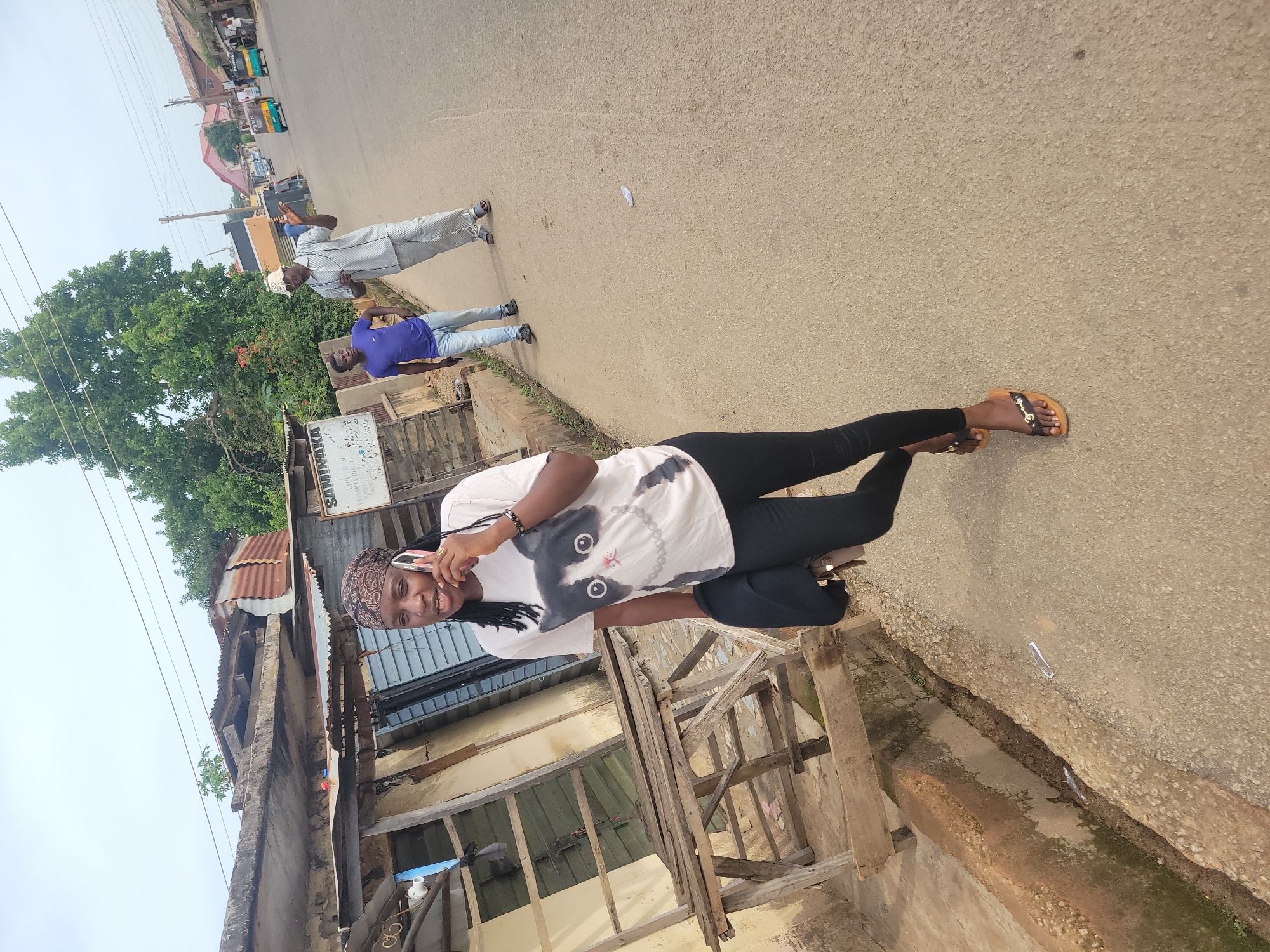 The image depicts a street scene in Jos, Nigeria. In the foreground, a young woman with braided hair and a headscarf is walking and talking on a pink smartphone. She is wearing a white t-shirt with a black and white cat graphic and black leggings, and brown sandals. She is carrying a dark bag over her left shoulder.
Behind her, on the right side of the street, two men are walking. One man is wearing a purple t-shirt and blue jeans. The other man is wearing a light-colored, striped outfit and a straw hat, and appears to be gesturing with his hands as he speaks.
The environment includes a paved road, a dirt shoulder with a concrete drainage ditch, and various buildings. Some of the buildings appear to be older structures with corrugated metal roofs and wooden elements, suggesting a less developed urban area. In the background, more buildings, trees, and a few vehicles, including what appear to be three-wheeled auto-rickshaws (Keke Napep), are visible, indicating a bustling street environment. The sky is overcast.