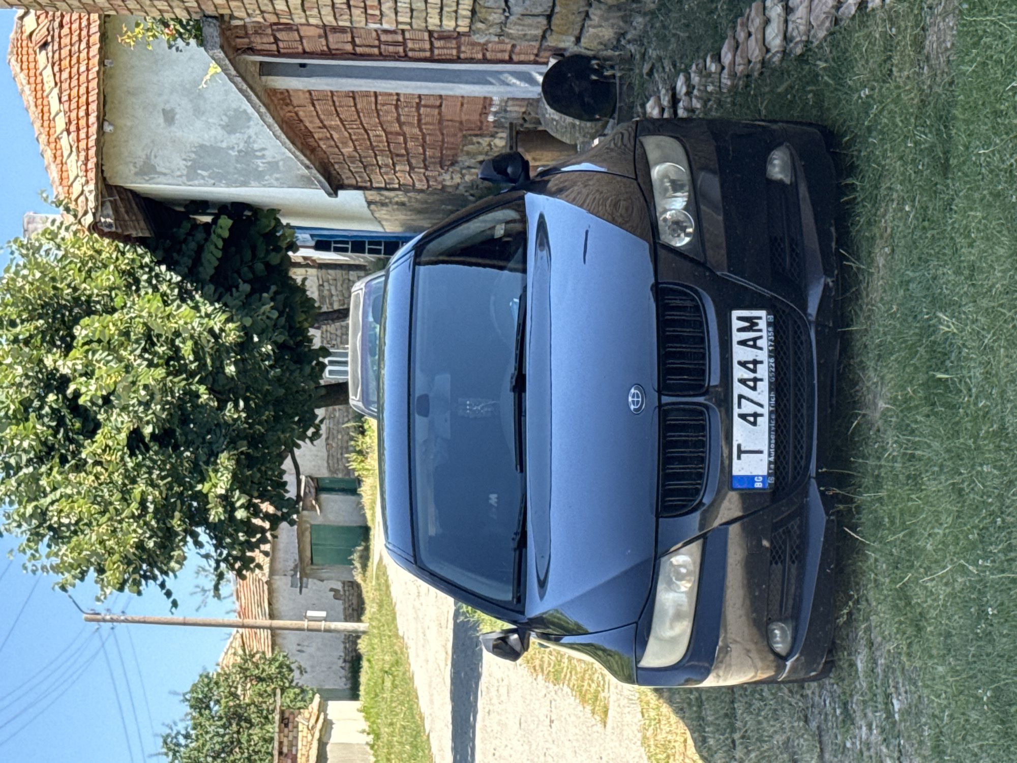 In Konop, Bulgaria, a black BMW sedan is parked on the side of a dirt road. The car is positioned in front of a narrow brick building and a lighter-colored structure with a red tile roof. A large tree with green foliage partially obscures the view of the buildings. The ground in front of the car is covered with green grass and some scattered stones, forming a low border along the road. The license plate of the car reads "T 4744 AM" with "BG" to the left and smaller text below. The sky is clear and blue.