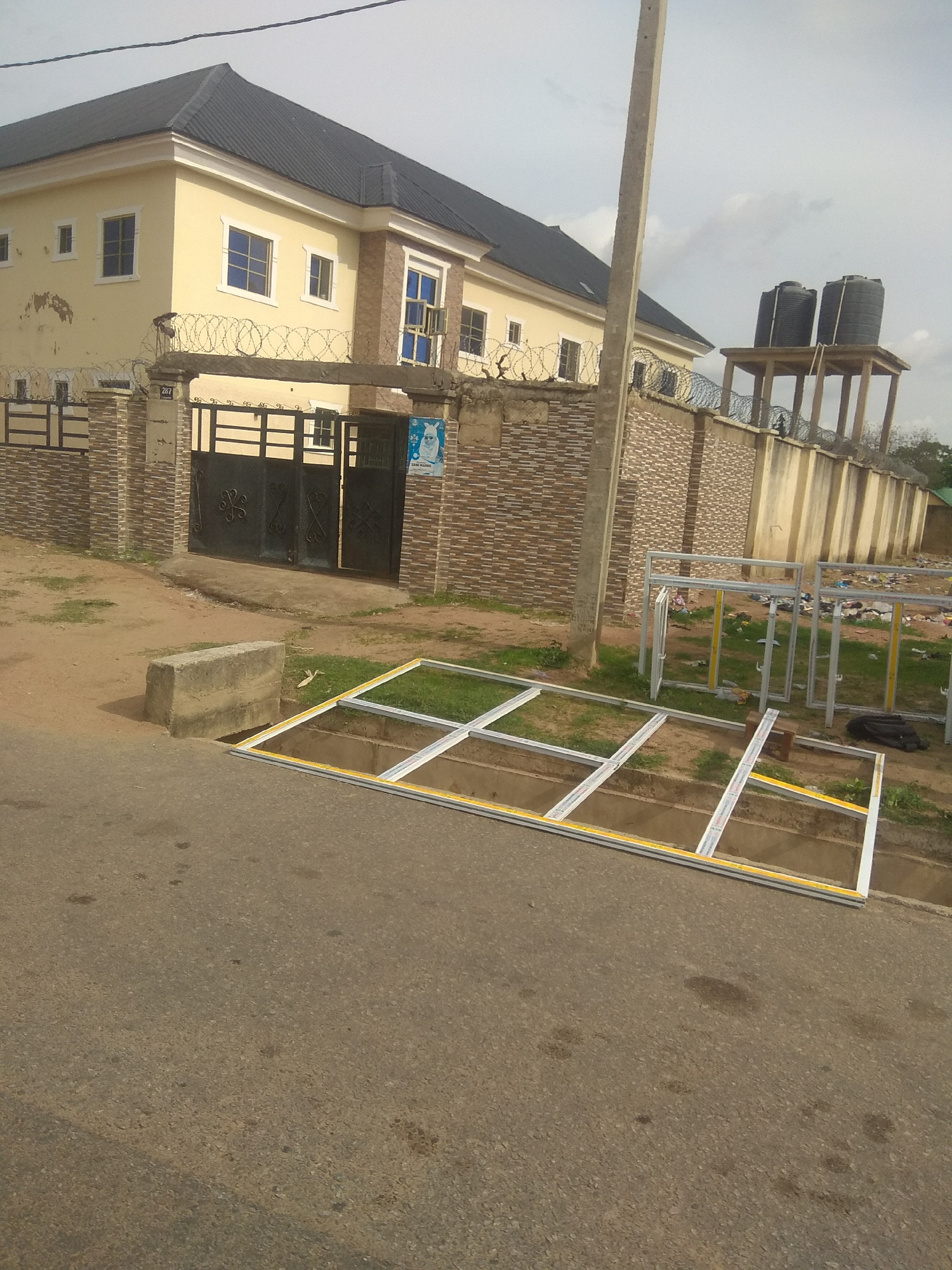 The image depicts a street scene in Jalingo, Nigeria. In the background, a two-story yellow building with a dark roof stands behind a brick wall topped with barbed wire. The building has several windows with blue panes. To the right of the building, two large black water tanks are elevated on a wooden platform. In the foreground, an unfinished metal frame, possibly for windows or doors, lies on the ground, partially in a ditch next to the asphalt road. Several other similar frames are visible further back, leaning against a wall and scattered on the grassy area. A concrete block is also present near the ditch. The sky is overcast.