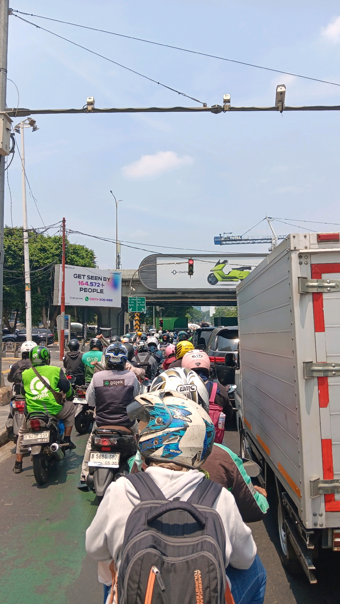 This is an eye-level, medium shot of a traffic jam in South Jakarta, Indonesia. The foreground is dominated by several motorcyclists, most wearing helmets and brightly colored jackets, indicating they may be delivery drivers. The middle ground shows a dense flow of motorcycles and some cars, all stopped in traffic. A large white truck is on the right side of the frame, partially obscuring the view of the traffic. In the background, there is an elevated road, a billboard advertising "GET SEEN BY 164,572+ PEOPLE," and a traffic light that is red. The sky is clear and blue with some scattered clouds, and there are power lines overhead. The scene appears to be on a sunny day.