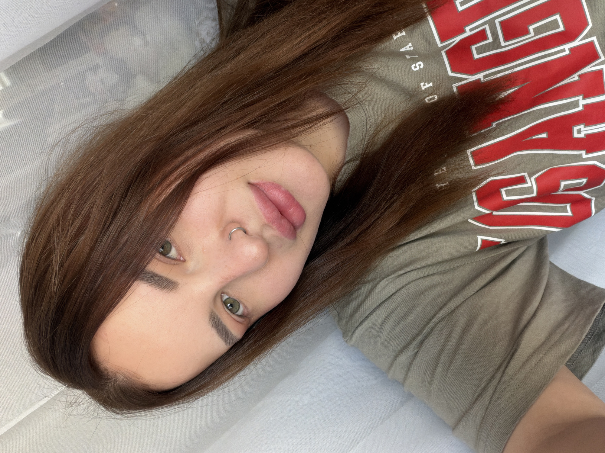 A close-up, angled, indoor shot depicts a fair-skinned person with brown hair and green eyes. They have a silver nose ring on their left nostril. They are wearing a gray t-shirt with a red and white logo. The background is a soft, white fabric. The location is Unknown, Unknown.
