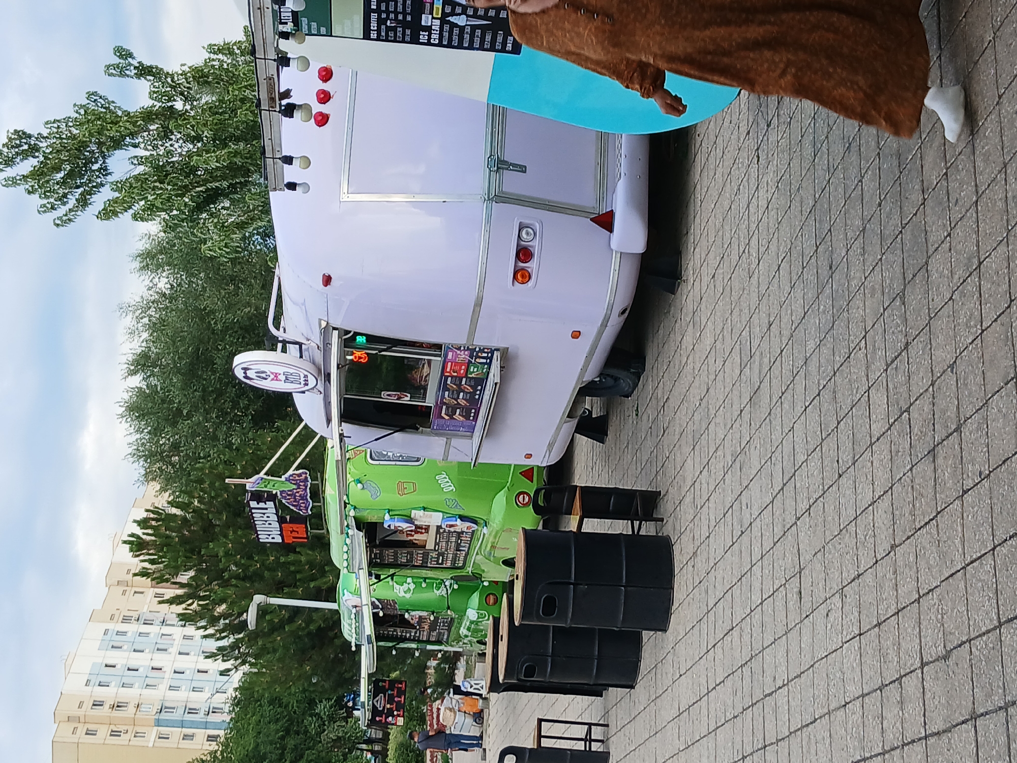 The image shows a row of food trucks parked on a paved area, likely an outdoor market or event. In the foreground, there are several black barrels, some stacked, serving as seating or decorative elements. Behind the barrels, a bright green food truck with colorful graphics is visible, advertising "BUBBLE TEA". Further back, a pale purple food truck is parked, its serving window open. A person wearing a long, brown, draped garment and a white sock is partially visible near the purple truck, appearing to interact with it. The background features lush green trees and a tall, multi-story building with many windows. The sky is overcast, suggesting a cloudy day. The overall scene appears to be a casual outdoor gathering, possibly during the daytime. Visible text includes "BUBBLE TEA" on the green truck and menu items on the serving windows of both trucks, though the text is not fully legible.