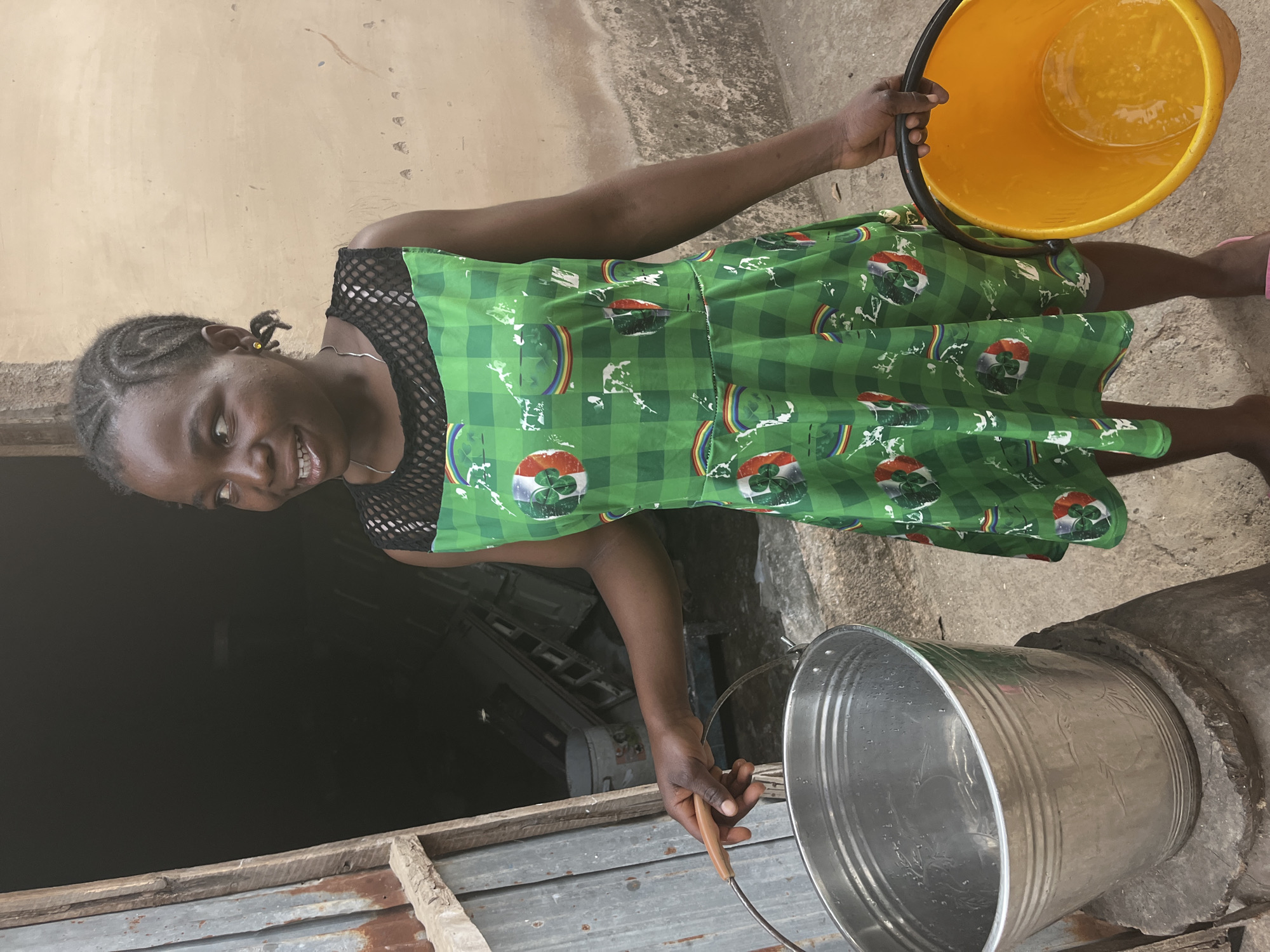 A young person, likely female, is depicted in an outdoor setting in Jos, Nigeria, engaged in the activity of water fetching. She has dark skin and her dark hair is styled in braids. She is wearing a vibrant green dress with a white and green checkered pattern, adorned with circular emblems featuring a rainbow and what appear to be shamrocks or clovers. A black mesh top is worn underneath the dress. A small gold stud earring is visible in her left ear, and she wears a simple silver necklace. She is looking towards the viewer with a gentle smile.
In her right hand, she holds a yellow plastic bucket containing water. Her left hand is positioned near a silver metallic bucket, also holding water, which rests on a concrete surface. The silver bucket has a simple wooden handle and some etched decorative patterns on its side. This bucket is situated next to a dark, shadowy opening, possibly indicating a well or a water storage area, framed by weathered wooden and metal structures.
The background consists of a light brown or tan wall on one side and the dark, recessed opening on the other. The ground appears to be concrete or packed earth. The scene is brightly lit, suggesting it is daytime. No visible text is discernible in the image. The overall impression conveys a moment of daily life involving water collection in a local community.