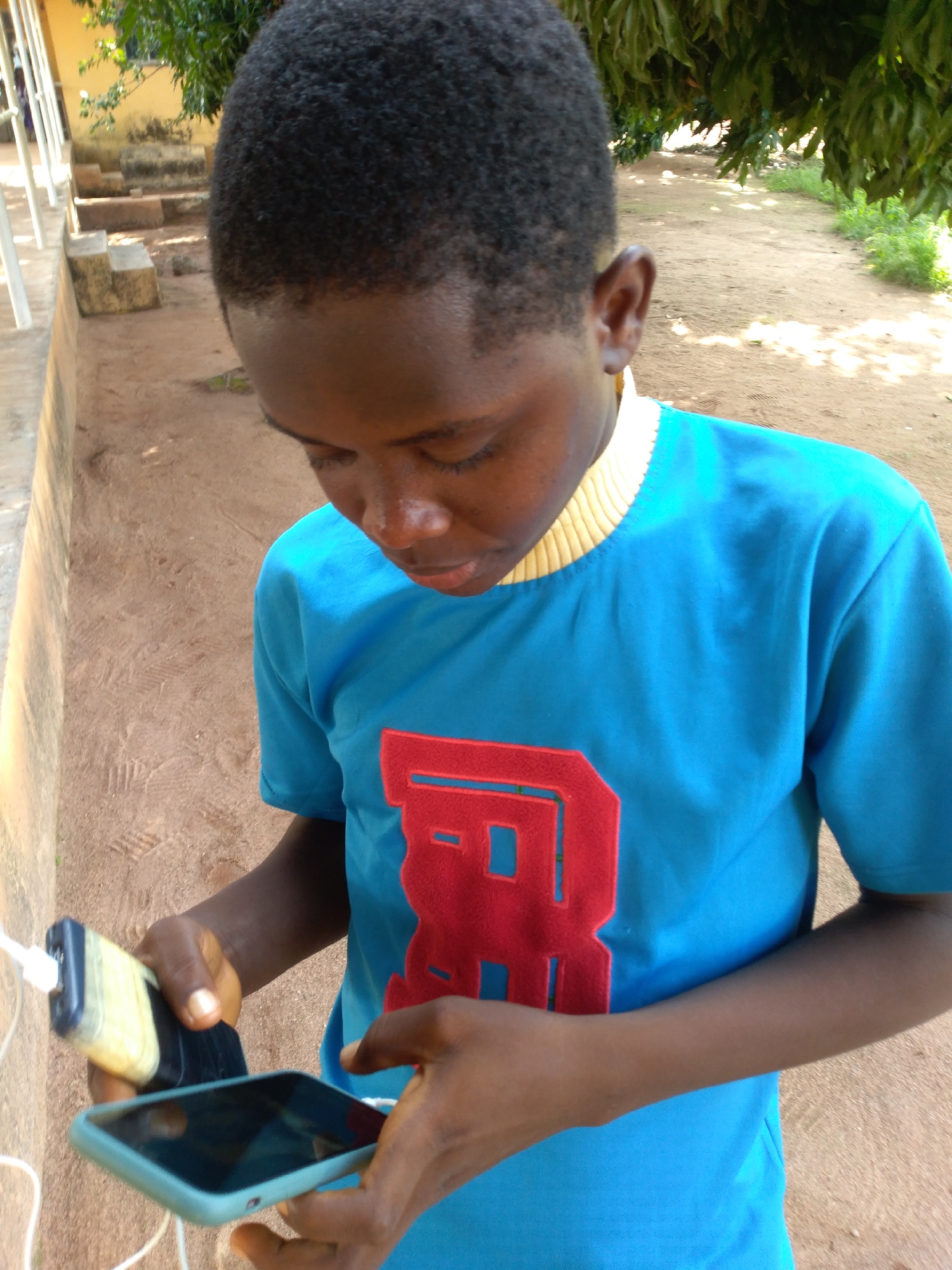 An individual with dark skin and short, curly dark hair is depicted outdoors in Jos, Nigeria, looking downward. The person is wearing a short-sleeved, light blue t-shirt featuring a prominent red letter "R" on the chest and a ribbed yellow collar. In their hands, the individual holds a light blue/green smartphone and a black and yellow portable power bank, which are connected by a white charging cable. The immediate environment consists of reddish-brown dirt ground with visible footprints. To the left, a light-colored concrete wall or ledge is present. The background features lush green foliage from trees, with sunlight dappling through the leaves, and an unpaved path visible beneath the trees.