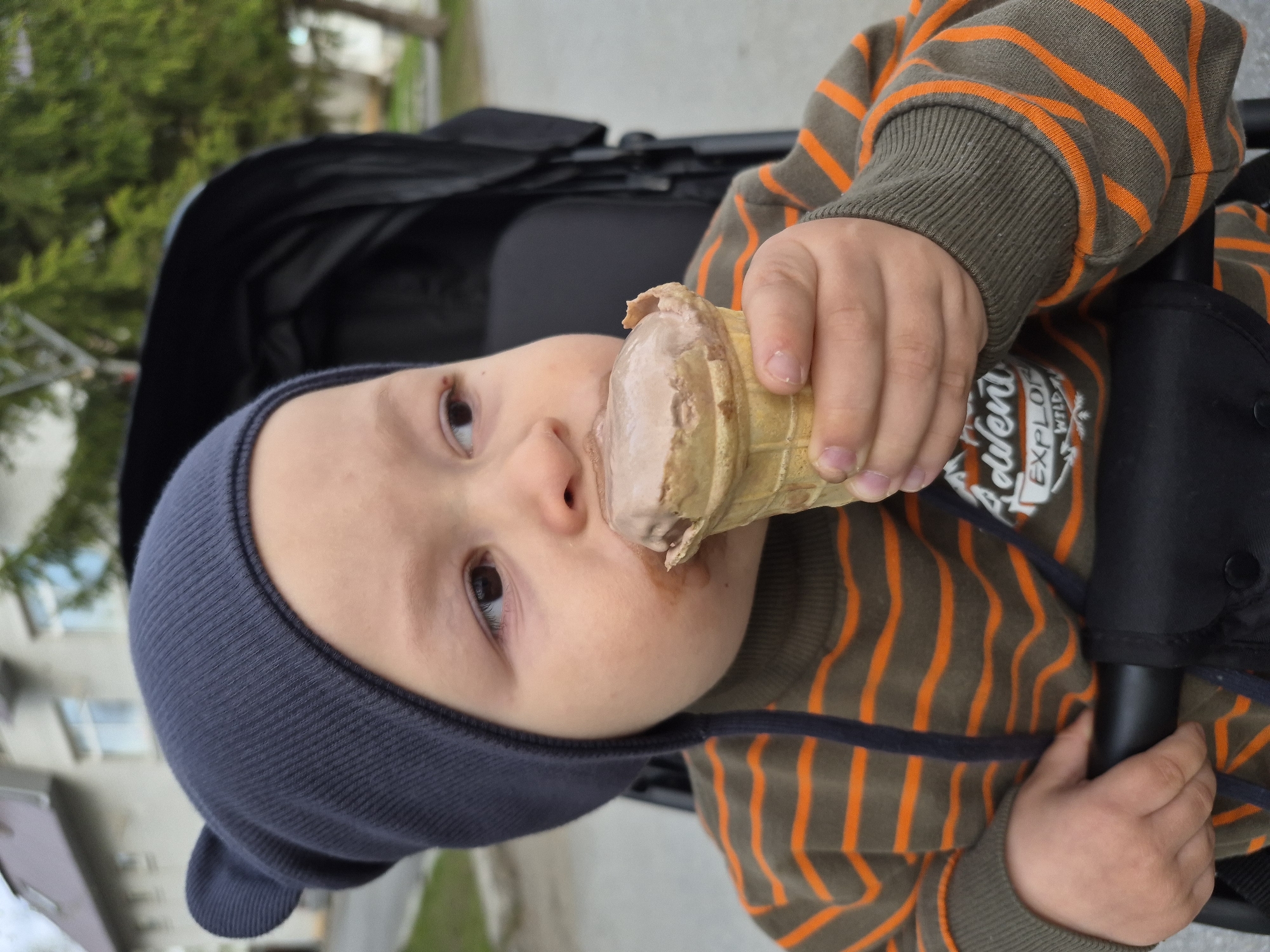 A young child in a stroller is eating an ice cream cone. The child is wearing a dark blue knit hat and a brown and orange striped shirt. The stroller is black. The background is blurred, but it appears to be an outdoor setting in a city with buildings and trees. The location is Tobolsk, Russia.