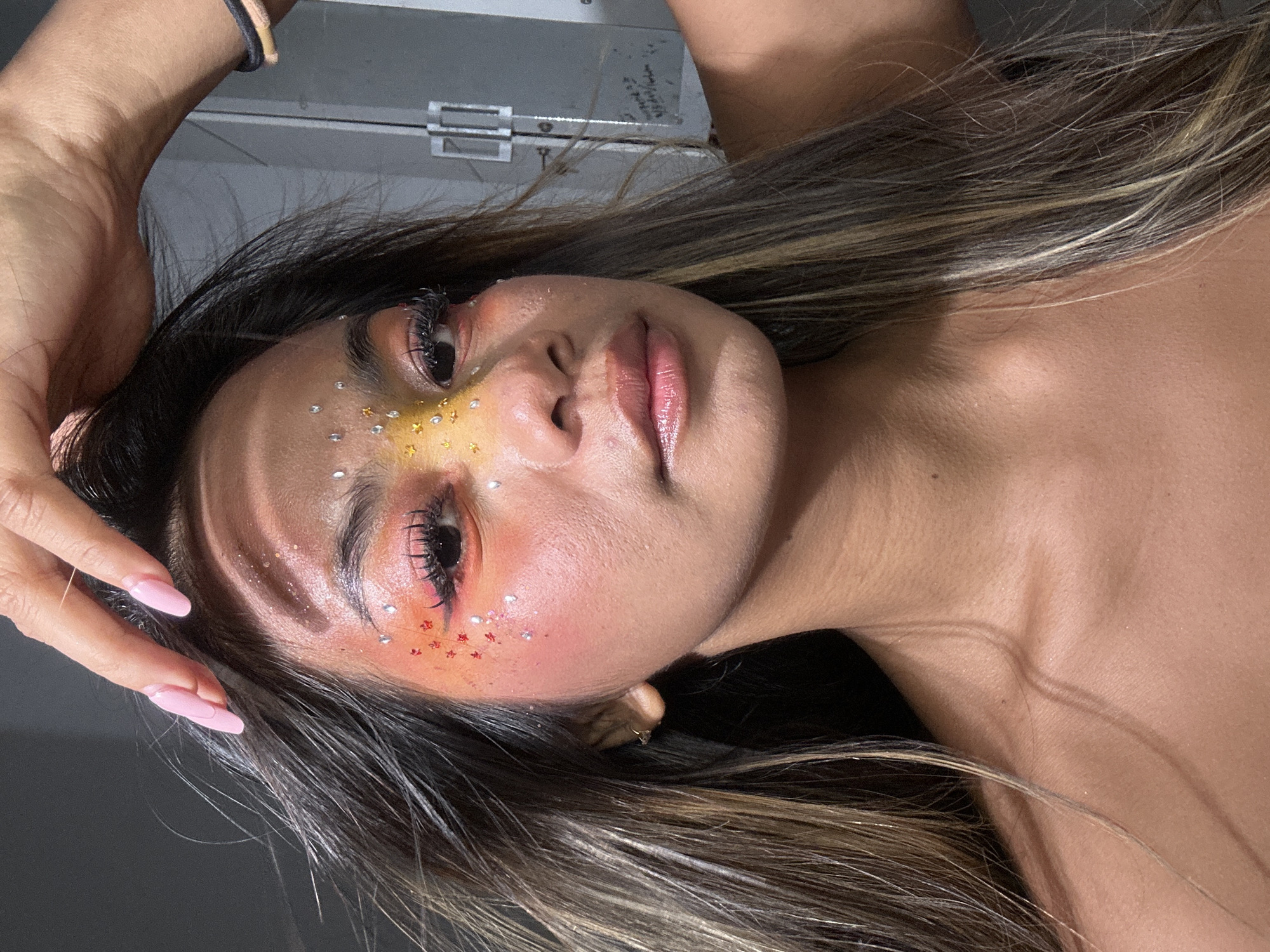 A young woman is lying down, facing the camera at an angle. She has brown hair with blonde highlights, and her skin is tan. She has elaborate makeup on her face. There are yellow and gold star-shaped sequins near her right eye and silver, gem-like sequins on her forehead. Her cheeks have a reddish-orange hue, and there are small, red star-shaped sequins around her left eye. Her lips are pink. Her left hand is raised, with her fingers near her face. Her nails are painted light pink. She is wearing a black elastic bracelet on her wrist. The background is mostly out of focus. It is unclear if the photo was taken in North Jakarta, Indonesia; there is no way to verify this using this image.