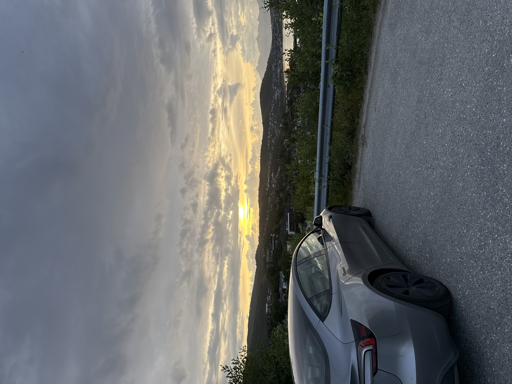 A silver electric car is parked on the right side of a paved road overlooking a city and a body of water at sunset. The car is angled towards the left, with its rear towards the viewer. The car's windows reflect the sky and surrounding landscape. The road curves to the left in the foreground. A metal guardrail runs along the edge of the road, separating it from a grassy embankment. Beyond the embankment, a city with houses is nestled amongst rolling hills. In the distance, a body of water is visible, and mountains can be seen on the horizon. The sky is filled with dramatic clouds, with rays of sunlight breaking through to illuminate the scene. The overall impression is one of a peaceful, scenic overlook. The location is likely on a hill above Harstad, Norway, offering a view of the city and its surrounding natural beauty during the evening hours.