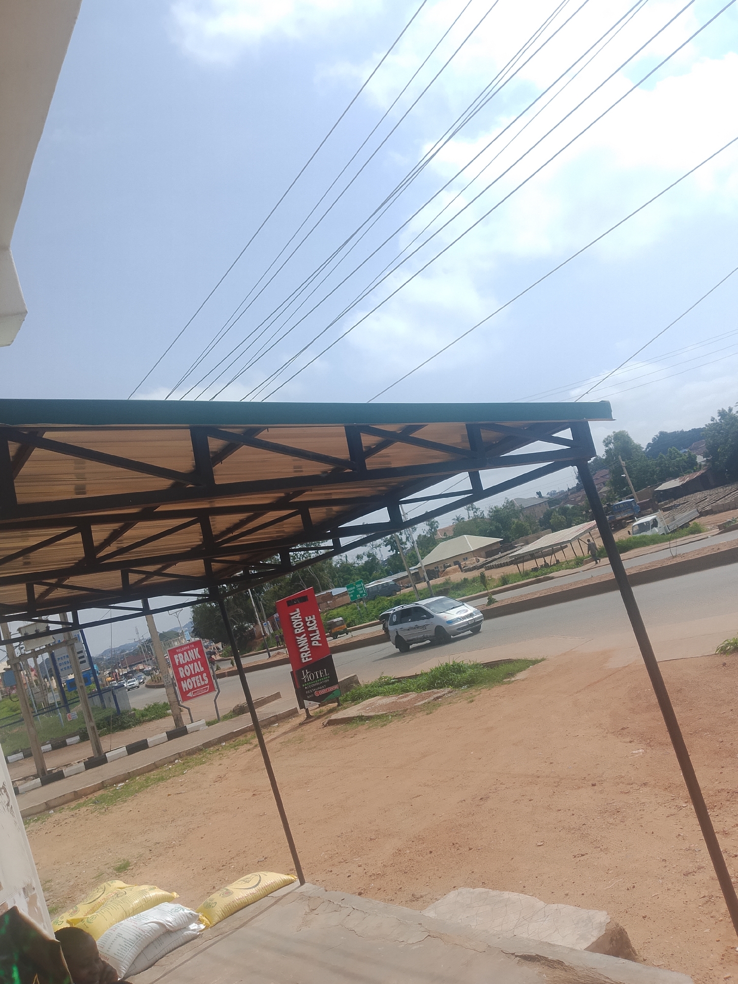 The image depicts an outdoor scene in what appears to be a roadside setting in Bukuru, Nigeria. A covered structure, likely a shelter or a stall with a corrugated roof supported by dark metal beams, dominates the foreground. Underneath the structure, there are bags of what looks like grain or fertilizer, suggesting a commercial or storage area.
To the left of the structure, there is a sign that reads "FRANK ROYAL HOTELS" and another sign with text that is difficult to decipher but appears to be in English. To the right, a red sign with white lettering states "FRANK ROYAL PALACE" and below it, "HOTEL", "RESTAURANT", and "BAR". These signs indicate the presence of hospitality businesses.
The scene includes a paved road with some traffic. A silver car is visible on the road. The background shows a mix of buildings, trees, and vegetation, indicative of a semi-urban or rural environment. The sky is partly cloudy, suggesting it is daytime. The overall atmosphere is that of a typical day in a Nigerian town, with elements of commerce and daily life. There are no people clearly visible engaged in any specific activity, but the presence of the road and businesses implies ongoing activity. The ground is a reddish-brown soil. The image is shot from a slightly elevated perspective, looking towards the road and beyond.