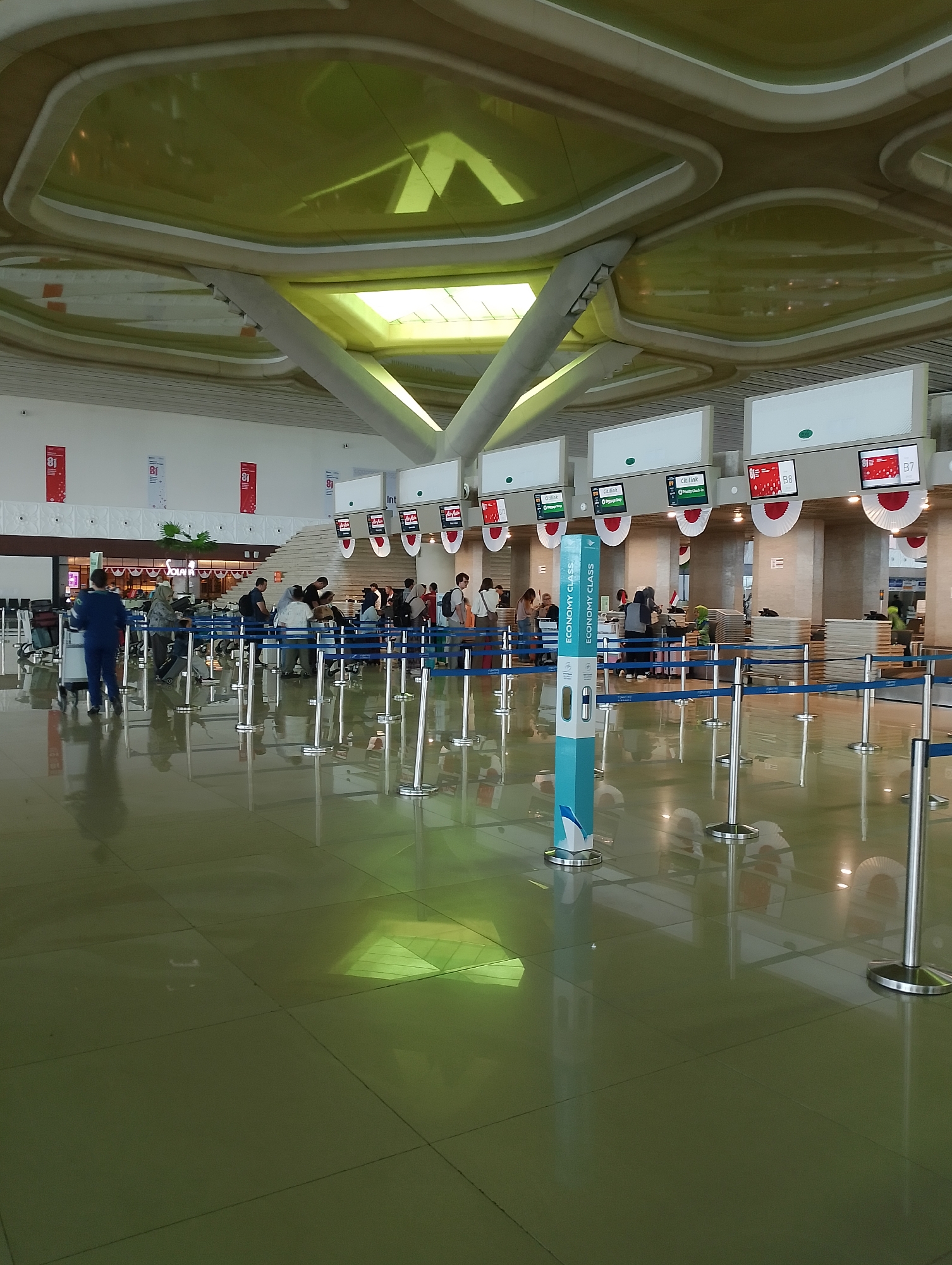 The image depicts the interior of an airport check-in area, likely at Yogyakarta International Airport in Palihan, Indonesia, given the presence of "Citilink" branding visible on some screens. The ceiling features an unusual, segmented design with yellow translucent panels and white ribbed framing, allowing light to filter through. Several check-in counters are visible, each equipped with digital screens displaying flight information. Passengers are lining up at these counters, with some interacting with staff. Blue retractable belt barriers are used to organize the queues. A prominent blue and white stand with "ECONOMY CLASS" branding is positioned in the foreground, likely indicating a specific queue or information point. The floor is highly polished and reflective, showing distortions of the ceiling and overhead lights. Luggage trolleys are present near some passengers. The overall atmosphere appears to be that of a busy travel hub.