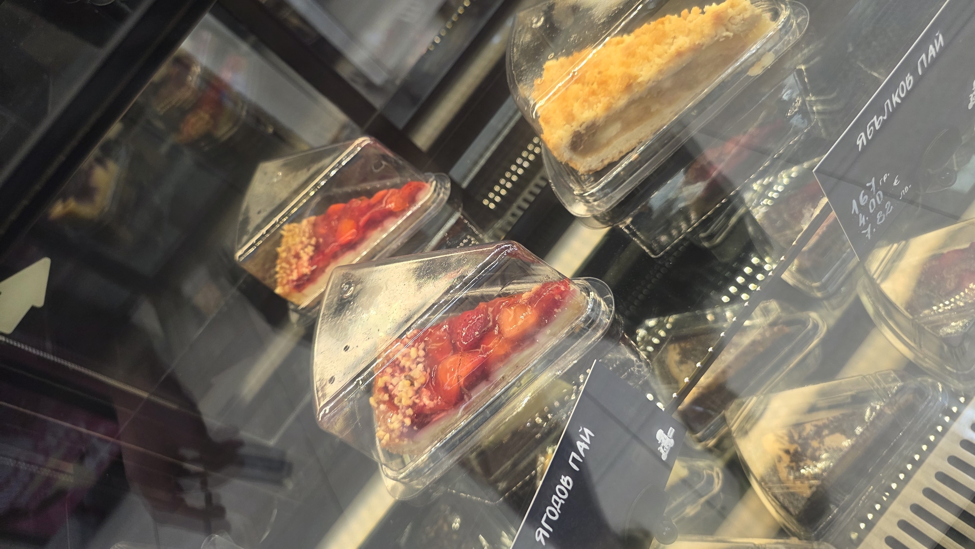 The image depicts a display case containing slices of pie in Burgas, Bulgaria. The pie slices are individually packaged in clear, triangular plastic containers. The pies include a strawberry pie, easily recognized by its bright red strawberry topping, and an apple pie, discernible by its golden-brown, crumble topping. Signage in Cyrillic is visible near one of the apple pies, indicating the name "ЯБЛОКОВ ПАЙ" (Apple Pie), along with prices in multiple currencies.