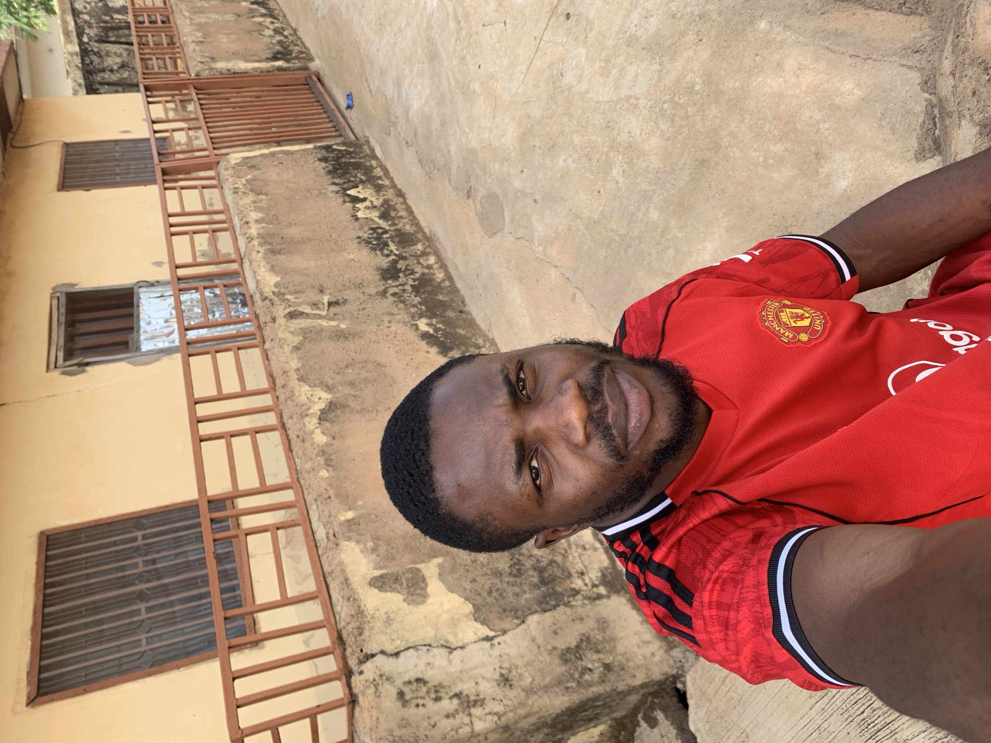 A man is taking a selfie while wearing a red Manchester United jersey. The background shows a weathered building with barred windows and a concrete surface, likely an exterior area. The location appears to be in an urban setting, potentially Rafin Par, Nigeria.