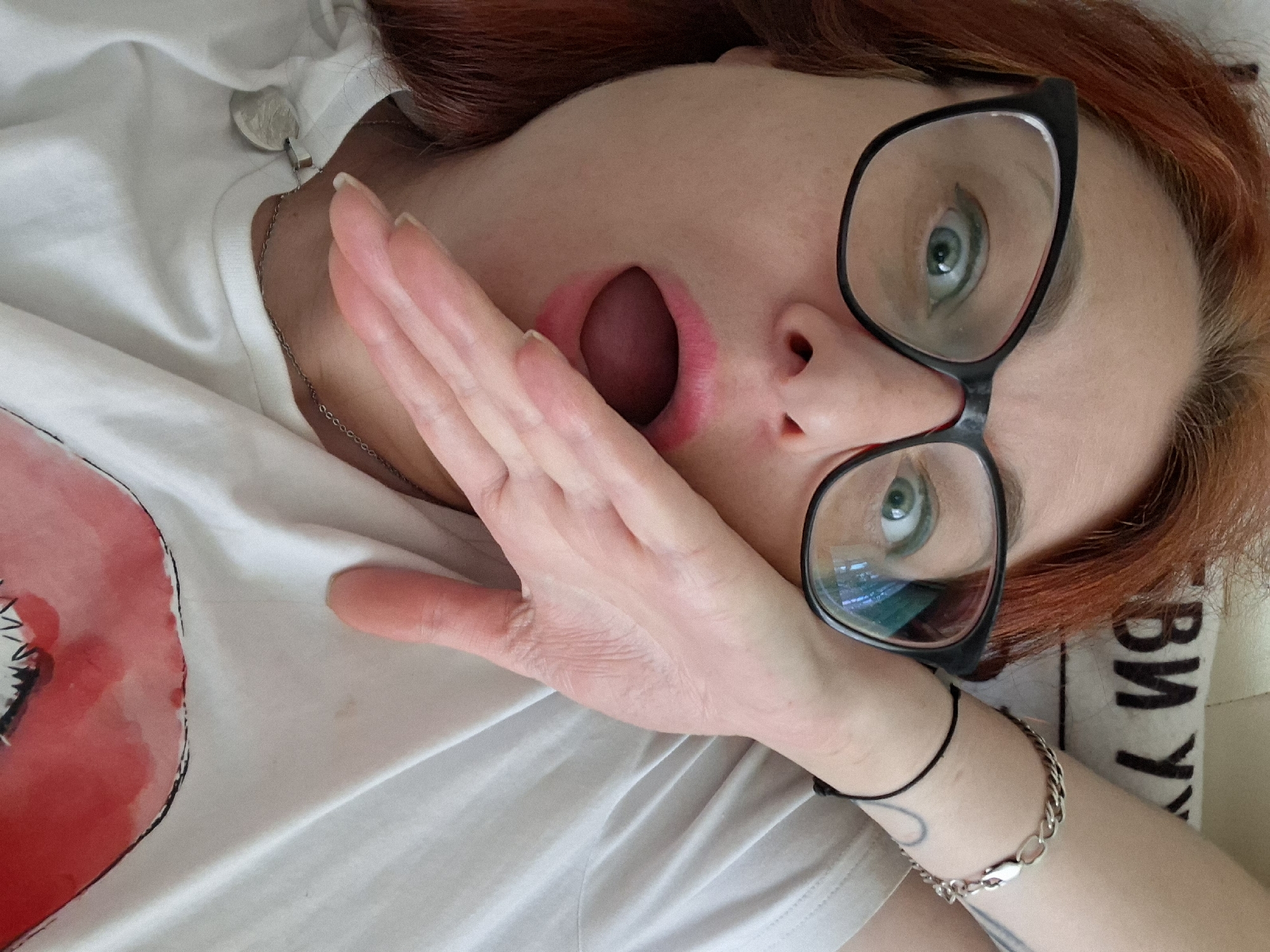 A medium shot captures a woman with auburn hair and glasses lying on her back and looking upwards. Her left hand is raised to her mouth, covering her open lips and pink lipstick. Her eyes are wide and her eyebrows are raised, suggesting surprise or shock. She is wearing a white t-shirt with a drawing of red lips on it. A silver chain necklace with a heart-shaped pendant is visible on her chest. On her right arm, a silver bracelet with a chain design is worn, along with a black thread bracelet. The background shows a light-colored surface with Cyrillic letters printed on it. There is no discernible urban setting or indication of Myadzyel, Belarus.