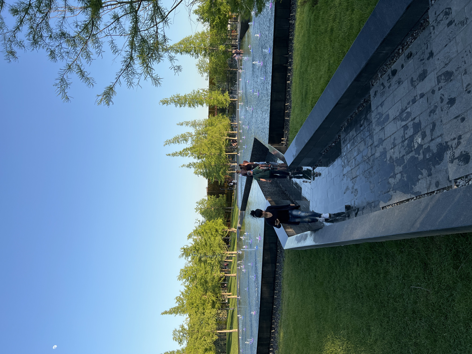 The image shows a scene in a park, likely the Krasnodar Park in Russia, featuring a long, narrow reflecting pool lined with trees. The pool is constructed with dark stone and includes small water jets. Several people are visible along the edge of the pool, some standing and others sitting on the stone border. The sky is a clear blue. Grass is present on the sides of the pool.
