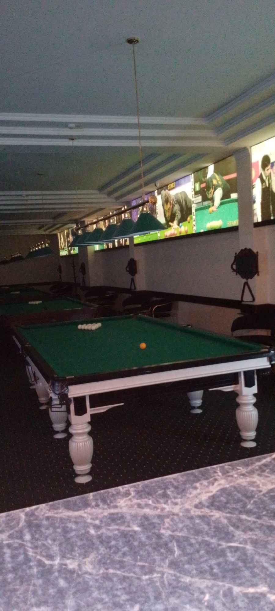 The image depicts an indoor billiards hall, possibly in Angren, Uzbekistan. In the foreground, a billiards table with a green felt surface is prominently displayed, featuring several white billiard balls arranged in a rack and one orange ball positioned to the right. The table has ornate white legs and trim. In the background, multiple other billiards tables are visible, some with their balls racked. The ceiling features decorative molding and is illuminated by overhead green lamps, typical for such establishments. Along the back wall, large screens display images of people playing billiards. The overall ambiance suggests a recreational or entertainment venue.