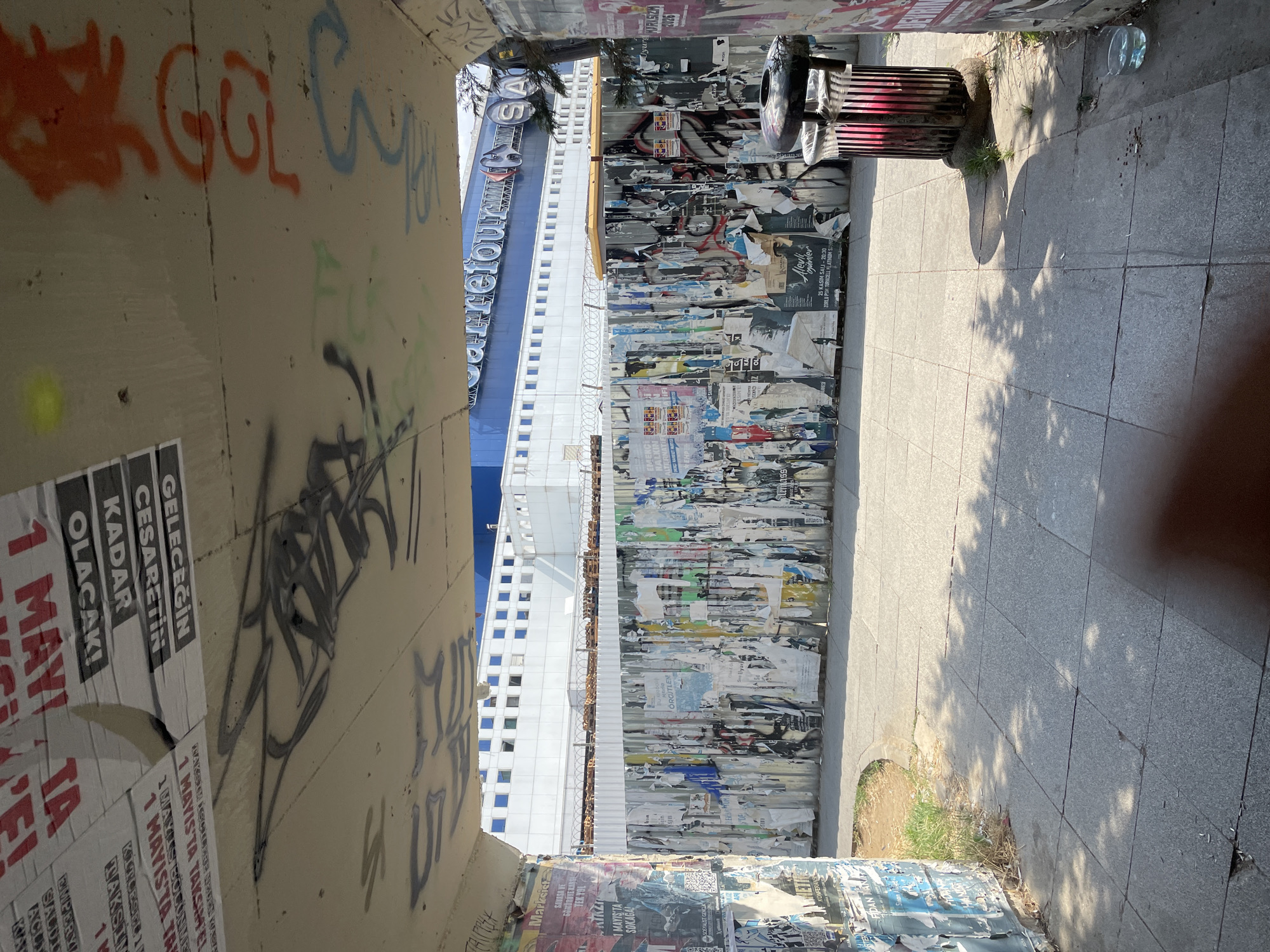The image captures a street scene in Kadıköy, Turkey. In the foreground, a paved walkway is visible, with dappled sunlight and shadows cast by overhead foliage. To the left, a wall is covered in layers of torn posters, some with visible text and images. To the right, a metal trash bin with a red interior stands on a stone base.
Further back, a corrugated metal fence, also heavily adorned with torn posters, stretches horizontally across the scene. Behind the fence, a large, modern building with a blue facade and numerous windows is visible, bearing the "CarrefourSA" logo. The sky above is bright and clear. The overall impression is one of an urban environment with visible layers of advertising and decay.