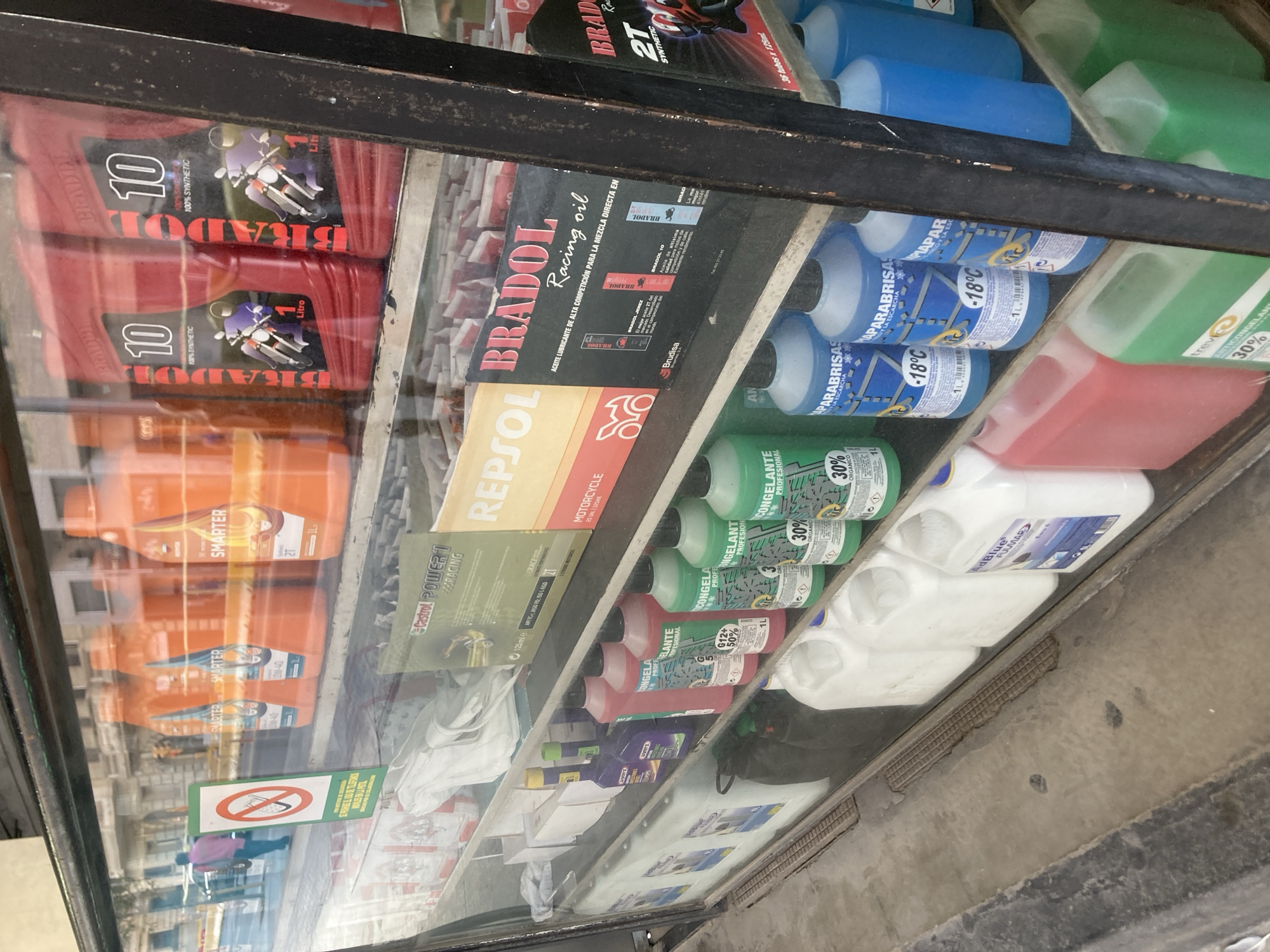 This is a photograph of a display shelf in what appears to be an automotive or motorcycle supply store. The shelves are filled with various liquids in plastic bottles and containers, primarily engine oils and antifreeze.
Prominent brands visible include "BRADOL," "REPSOL," and "Castrol." Some bottles are red, blue, and green, with labels indicating different viscosities, types of oil (e.g., "100% SYNTHETIC," "2T"), and intended uses (e.g., "Racing oil," "MOTORCYCLE"). There are also bottles labeled "PARABRISAS -18°C" which suggests windshield washer fluid designed for cold temperatures. A container labeled "AdBlue" is also visible, commonly used in diesel vehicles.
In the foreground, a green rectangular sign with a red circle and a diagonal line over a cigarette indicates a "No Smoking" rule. The reflections in the glass display case show the street outside, with buildings that suggest an urban setting, possibly in Barcelona as indicated by the location context. The overall impression is of a well-stocked retail environment. No people are clearly depicted within the display, though figures can be seen through the glass in the street reflection. The lighting suggests it could be daytime.