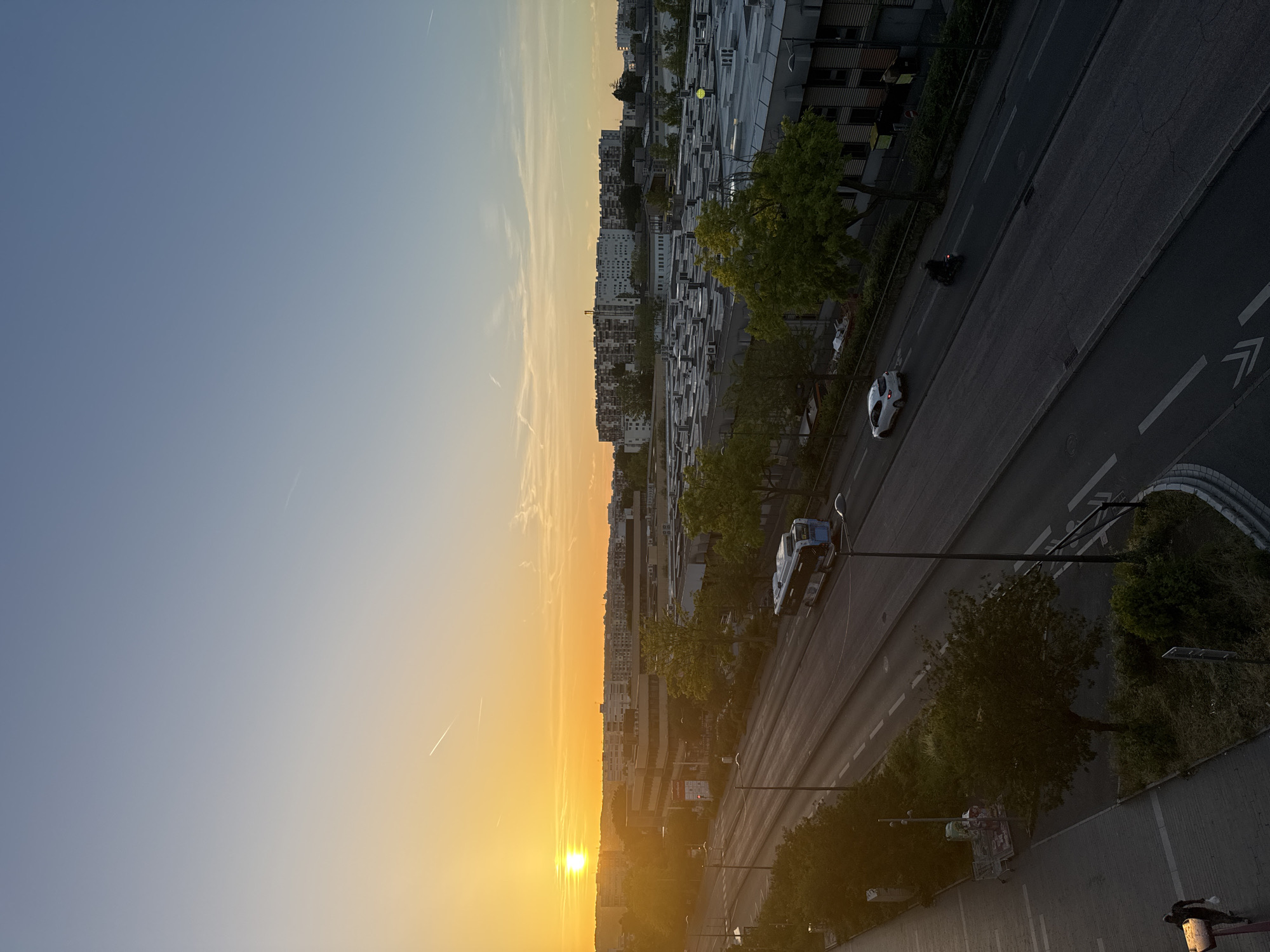 The image shows a sunset over a city. The sun is a bright orange orb low on the horizon, casting a warm glow across the sky and silhouetting buildings and trees in the foreground. A street runs diagonally through the scene, with a bus and a car visible. In the distance, a cluster of apartment buildings forms a skyline. The sky is clear with a few wispy clouds. The overall mood is peaceful and serene, with the sunset creating a dramatic and beautiful atmosphere.