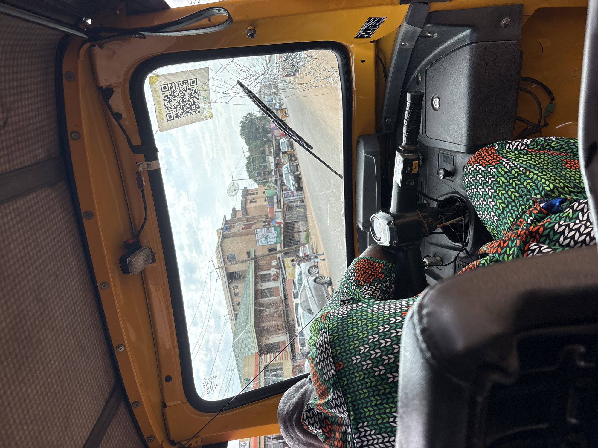 The image provides an interior view from a yellow auto-rickshaw, likely in Nkpor, Nigeria, showing the windshield and dashboard. A person, wearing a patterned green, white, and orange fabric, is seated in the driver's position, their back to the camera, operating the vehicle. The windshield is cracked. Outside the vehicle, a street scene unfolds with buildings lining the road, several parked cars, and other vehicles, including at least one other auto-rickshaw, in motion. The sky is overcast with scattered clouds. A QR code sticker is affixed to the upper right portion of the windshield.