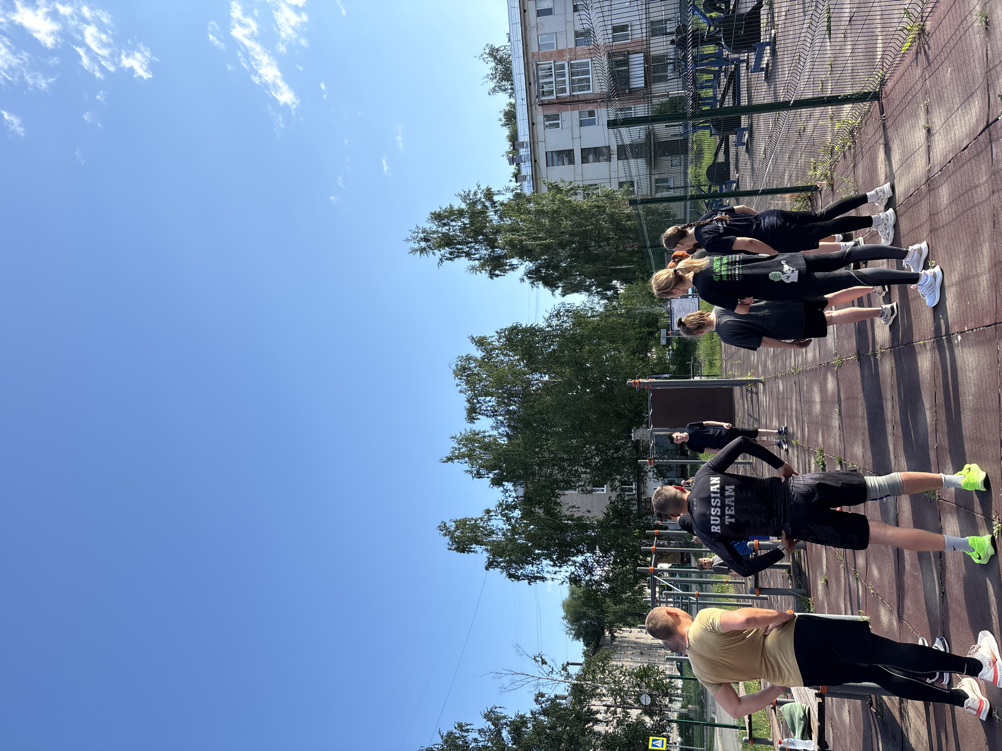 This media file captures an outdoor fitness session featuring a group of individuals, primarily young adults, engaged in exercise in Chaikovsky, Russia. The scene is set in a dedicated outdoor workout area with a reddish-brown, possibly rubberized, ground, under a clear, bright blue sky with scattered white clouds, indicating a sunny day, likely midday or early afternoon given the strong shadows.
In the foreground, two men are prominently visible from behind. The man closest to the camera wears a tan t-shirt, black shorts, and black leggings, with a water bottle placed nearby. Directly behind him, another man wears a black long-sleeved athletic top with the words "RUSSIAN TEAM" printed in white across the back. He also wears black shorts, grey leggings, and striking bright yellow-green sneakers. Both men adopt a rigid posture, hands on hips, suggesting a static exercise or attentive stance.
Further back, a line of women, also dressed in dark athletic wear, including t-shirts, leggings, and white sneakers, are visible, appearing to be in a similar exercise or formation. In total, at least eight people can be identified participating.
The background features various pieces of metal outdoor gym equipment, including pull-up and parallel bars, visible through a chain-link fence that borders the workout area. Beyond the fence, lush green trees separate the exercise space from several multi-story residential buildings, which form the backdrop. A yellow diamond road sign is partially visible in the bottom left corner, suggesting proximity to a public road. The overall atmosphere is one of focused physical activity in an urban outdoor setting.
