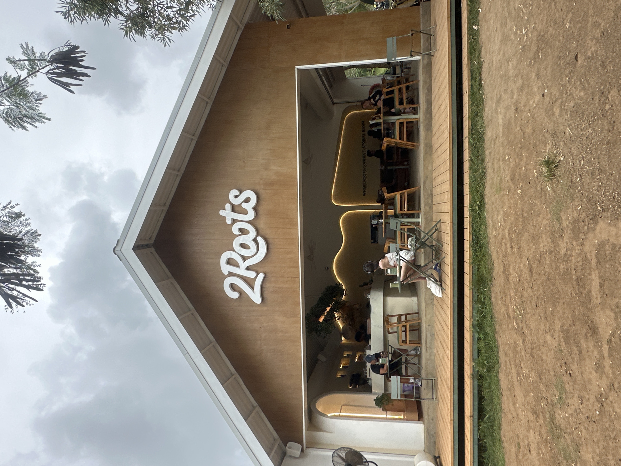 The image shows a facade of a cafe named "2Roots" in Kuta Selatan, Indonesia. The building has a pitched white roof and a wooden exterior. The main entrance is wide open, revealing the interior of the cafe. Inside, there is a counter and several tables and chairs where patrons are seated. A large, stylized plant adorns the interior. The cafe is situated on a wooden deck, which is surrounded by dry, brown ground and some patches of green grass. The sky is overcast. A person is seated at a table on the deck, facing away from the camera, seemingly engrossed in a book. Another person is seated at a table inside the cafe. In the foreground, on the deck, there are several empty chairs and tables. A pedestal fan is visible to the left of the entrance. A white bag is placed on the ground near the seated person on the deck. The overall scene suggests a casual outdoor dining or cafe environment.