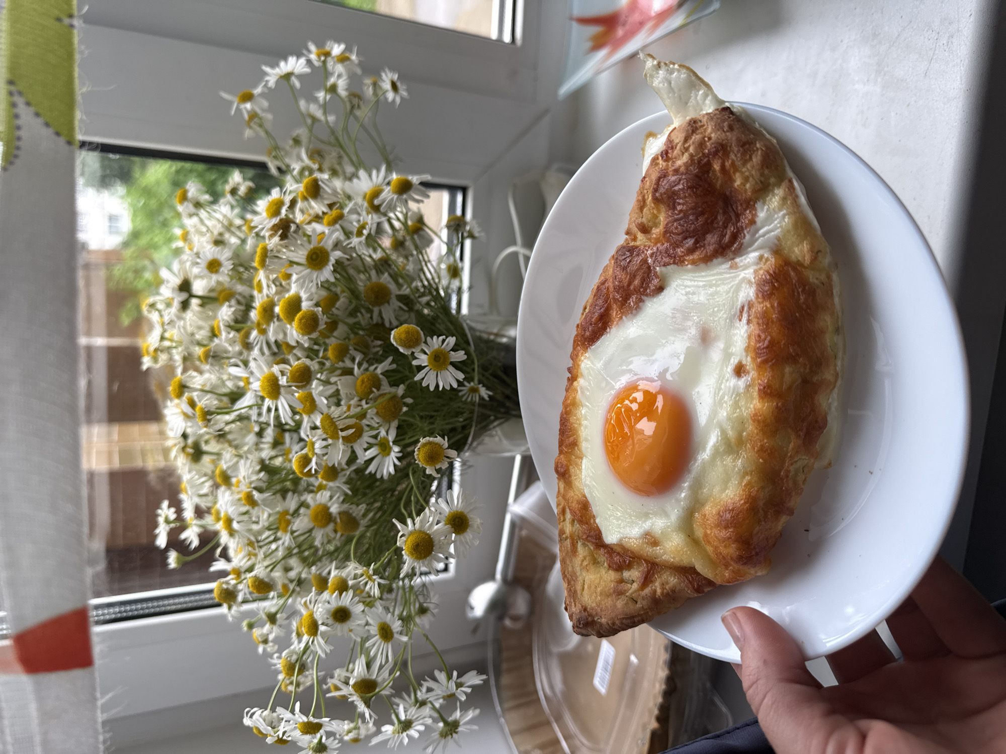 A close-up shot features a plate of khachapuri, a Georgian cheese-filled bread, topped with a fried egg. The bread is golden brown and appears to be slightly crispy. The egg has a bright orange yolk and fluffy white. In the background, a bouquet of daisies is arranged in a glass vase, with a window visible behind it. The overall scene suggests a casual meal setting, possibly at home, given the domestic elements like the window and curtains. The image appears to be taken indoors, near a window, as suggested by the natural light illuminating the scene. The location is identified as Chaikovsky, Russia.