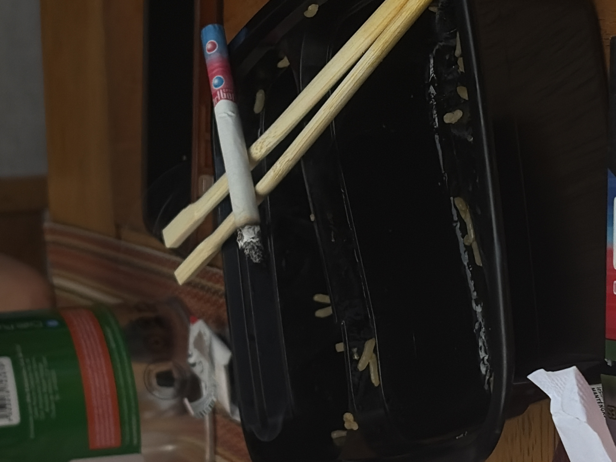 A partially smoked cigarette with a blue and red-striped filter rests atop a pair of wooden chopsticks inside a black, rectangular takeout container. The container exhibits food residue on its interior surface. A green and red can is partially visible in the lower-left corner of the frame. A white napkin lies to the right of the container.
There is no information available to determine whether this image was taken in the city of San Bernardino Tlaxcalancingo, Mexico.