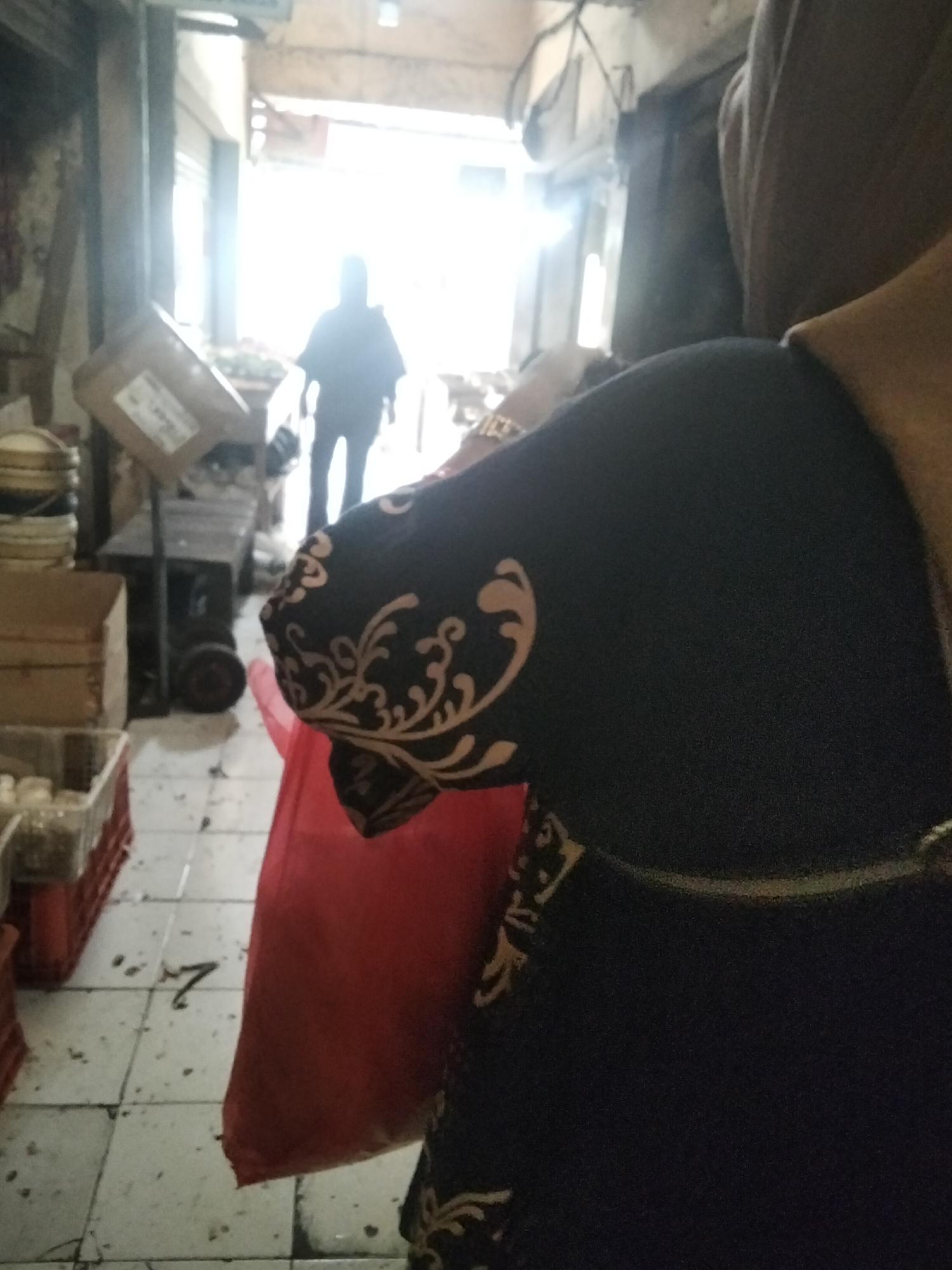 The image depicts an indoor market scene, possibly a traditional market, in Cisaat, Indonesia. In the foreground, a person wearing a dark headscarf and a dark garment with an intricate tan-colored floral pattern on the sleeves is partially visible, facing away from the camera. The person is holding a large red bag. The background is blurred and shows an aisle within the market, with sunlight streaming in from an opening at the far end. A silhouetted figure of a person is visible walking towards the light in the distance. Various market elements are present, including stacked crates, boxes, a trolley with wheels, and what appear to be containers or goods on shelves. The floor is tiled and appears somewhat dirty. The overall impression is that of a busy, everyday scene in a Southeast Asian market.