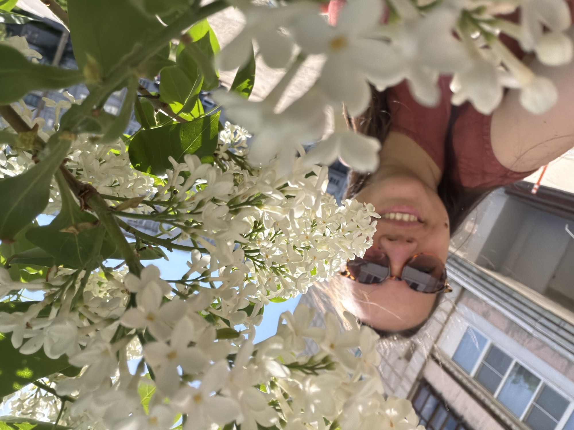 A woman with shoulder-length hair and sunglasses smiles into the camera. In the foreground, there are several branches of white lilac blossoms with green leaves. In the background, there is a building with multiple windows. The location is potentially Chaikovsky, Russia.