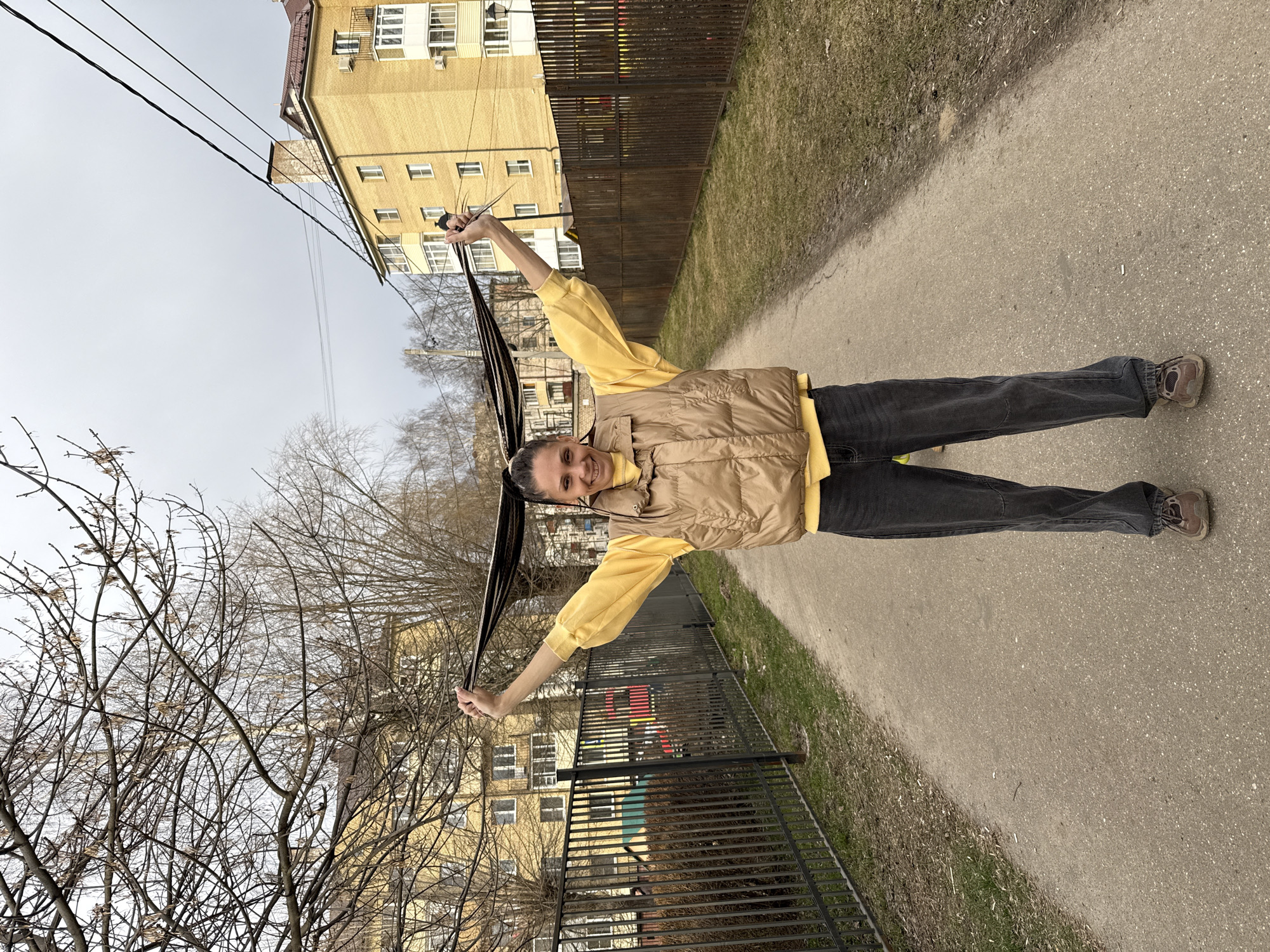 A woman stands smiling on a narrow paved path in Rusanovo, Russia, holding her long braids outstretched above her head. She wears a yellow long-sleeved shirt, a tan quilted vest, dark jeans, and brown shoes. A row of brown and green grass runs along one side of the path, and a black metal fence separates it from a grassy area and a multi-story apartment building with yellow brick and white framed windows. Bare tree branches are visible behind the fence. Power lines stretch across the top left corner of the frame against a cloudy sky.