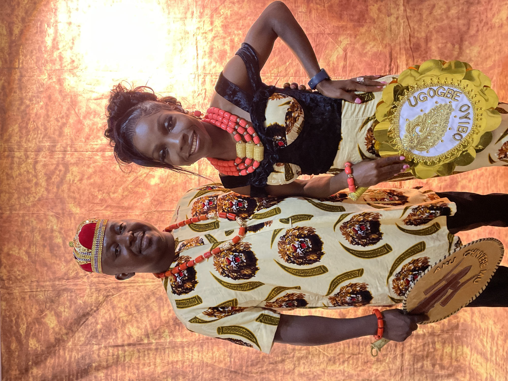 This portrait, taken in Barakin Akawo, Nigeria, features a man and a woman, both smiling warmly, dressed in elaborate traditional Nigerian attire. They are posing against a warm, mottled orange and brown backdrop, suggesting an indoor setting for a cultural event or celebration.
The man, on the left, wears a light cream-colored long-sleeved shirt patterned with brown lion heads and green horn-like motifs. He is adorned with a red and gold beaded crown and a multi-strand coral bead necklace, with a matching bracelet on his right wrist. He holds a light brown, oval-shaped traditional fan, which is carved with symbols and bears the text "ENWERU INU".
The woman, standing beside him, wears a black sleeveless top paired with a skirt made of fabric that matches the man's shirt. Her neck is adorned with a prominent, multi-layered coral bead necklace, complemented by coral bead bracelets on both wrists. Her hair is styled neatly, and she wears a wristwatch on her left arm. In her left hand, she holds a decorative golden circular piece with a white center, featuring intricate gold embroidery and the bold gold lettering "UGOGBE OYIBO". Both individuals display joyful expressions, contributing to the festive and celebratory ambiance of the scene.