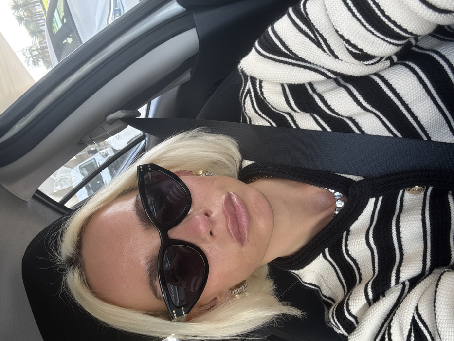 A woman with blond hair, wearing black sunglasses and a black and white striped sweater, is seated in a car. She has on a black seatbelt. The interior of the car is visible, along with a glimpse of the outside environment through the window. The image is a close-up selfie taken from a slightly elevated angle.