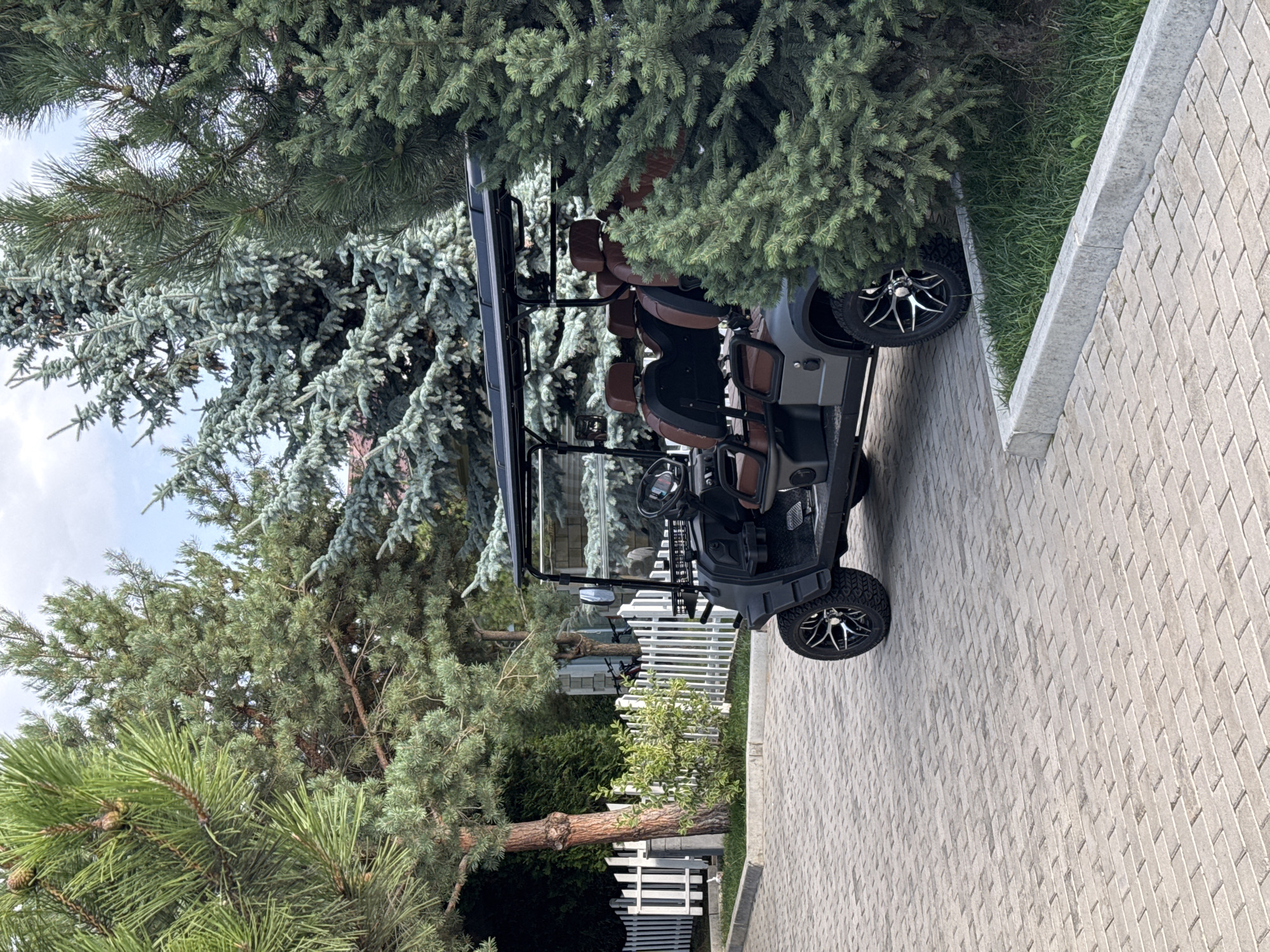 In Sary-Oy, Kyrgyzstan, a dark gray golf cart with brown leather seats is parked on a paved walkway lined with gray concrete borders and grass. The golf cart has a black roof and chrome-accented wheels. Behind the golf cart, a white picket fence separates the walkway from a grassy area with bushes and trees. Various types of evergreen trees, including pine and spruce, surround the scene. The sky is visible in the upper portion of the image, with some clouds present. The overall environment appears to be a well-maintained outdoor space, possibly a resort or residential area. No people are visible in the image.