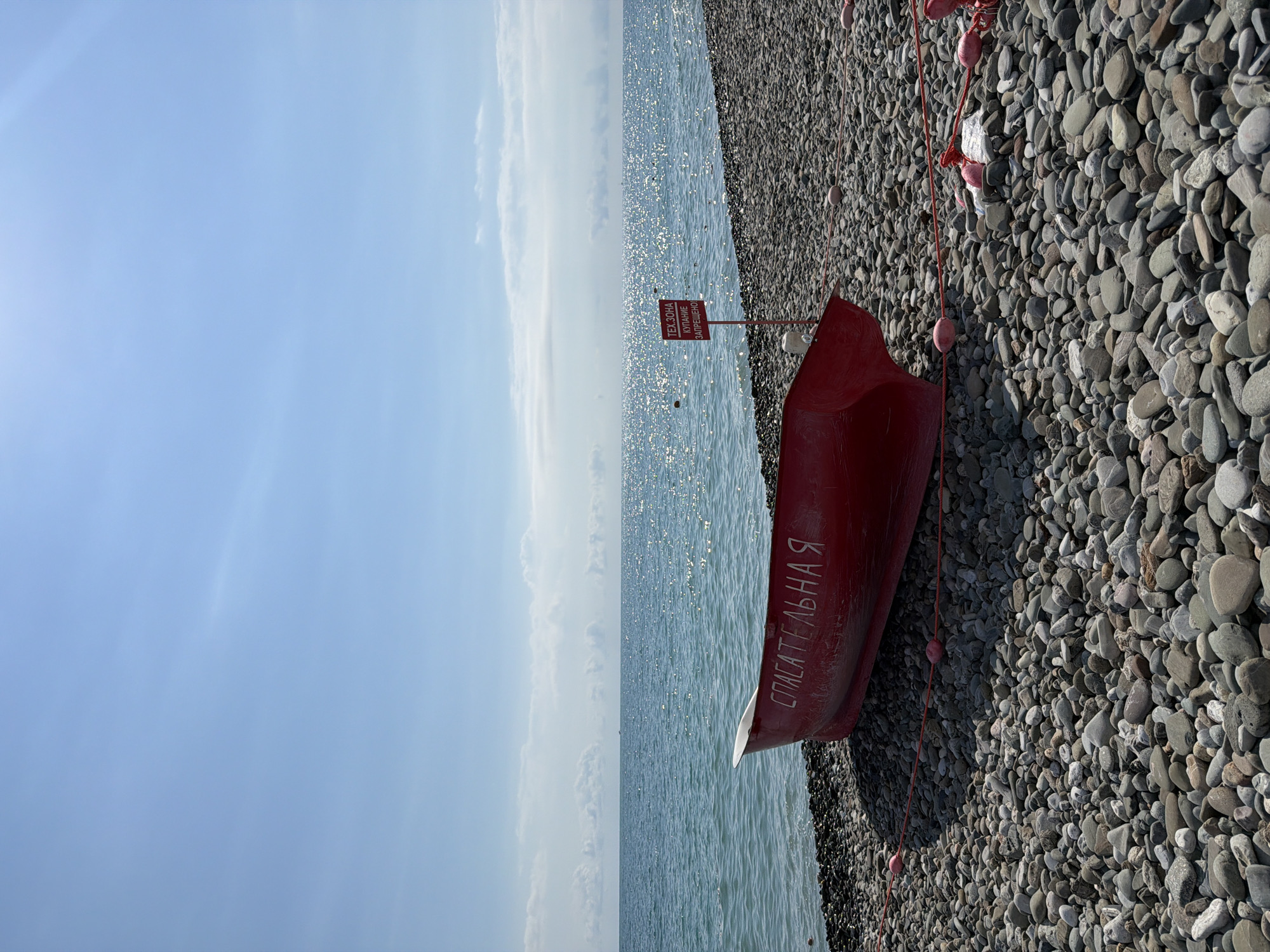 A bright red, weathered rowboat rests on a rocky beach near the shimmering, sun-drenched Black Sea in Sirius, Russia. The word "СПАСАТЕЛЬНАЯ" (meaning "rescue") is painted in white on the boat's hull. A small, red signpost with white lettering is planted in the pebbles near the water's edge, indicating a "ТЕХ. ЗОНА КУПАНИЕ ЗАПРЕЩЕНО" (technical zone, swimming prohibited). The sky is a clear, pale blue with wispy white clouds and the sun creates a dazzling reflection on the water. The beach is covered in smooth, grey, and brown pebbles. A red rope with pink buoys runs along the foreground, delineating the area. The scene is quiet and appears to be a still, sunny day at the seaside.