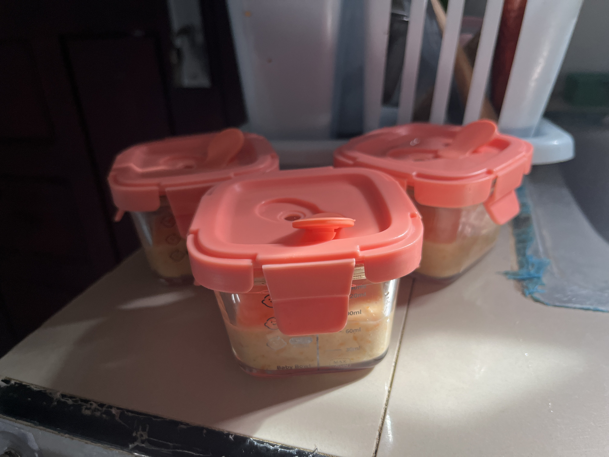 The image depicts three small, rectangular food storage containers with clear bodies and coral-pink, square-shaped lids, resting on a light-colored, tiled surface. Each lid is secured by four locking clips. The central container's lid features a prominent, raised, circular orange vent or seal button, while the lids of the containers on either side have a smaller, less distinct central flap or vent. All containers hold a light orange or yellow pureed substance, consistent with baby food, filled to approximately half to two-thirds capacity. The central container's clear body displays "Baby Bowl" text at the base, along with measurement markings (e.g., "20ml", "50ml", "MAX LOAD") and small stylized baby head icons printed on its side.
In the blurred background to the right, a white plastic dish drying rack is partially visible, holding what appear to be utensils. Behind the rack, a large, out-of-focus, light-colored object, possibly an appliance or cabinet, is present. A crumpled or textured blue cloth lies on the surface to the far right. The background to the left is dark and indistinct, suggesting an interior wall or furniture. The scene is illuminated by indoor ambient light, casting soft shadows beneath the containers. No individuals are present in the image.
There are no discernible visual elements within the image that allow for the identification of a specific geographic location, such as the city of Lhokseumawe, Indonesia.