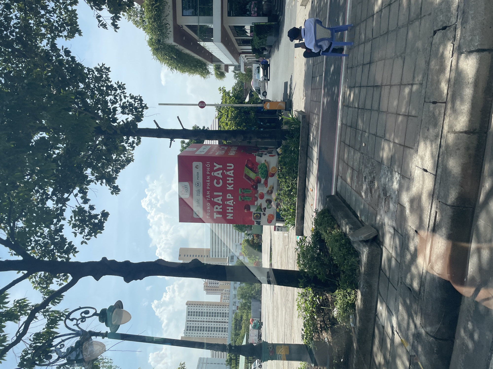 The image depicts a street scene in Thủ Đức City, Vietnam. On the right side of the frame, a man is seated on a blue plastic chair, looking at his mobile phone. He is wearing a white shirt and a dark cap. A large, red advertising billboard stands prominently in the middle of the scene, displaying images of various fruits and the text "TRÁI CÂY NHẬP KHẨU" (Imported Fruits). Behind the billboard, a modern building with lush greenery on its balconies can be seen. Trees line the street, providing shade. In the background, tall apartment buildings and a truck are visible. The sky is partly cloudy. The street is paved with concrete tiles, and there are planters with green foliage along the sidewalk.